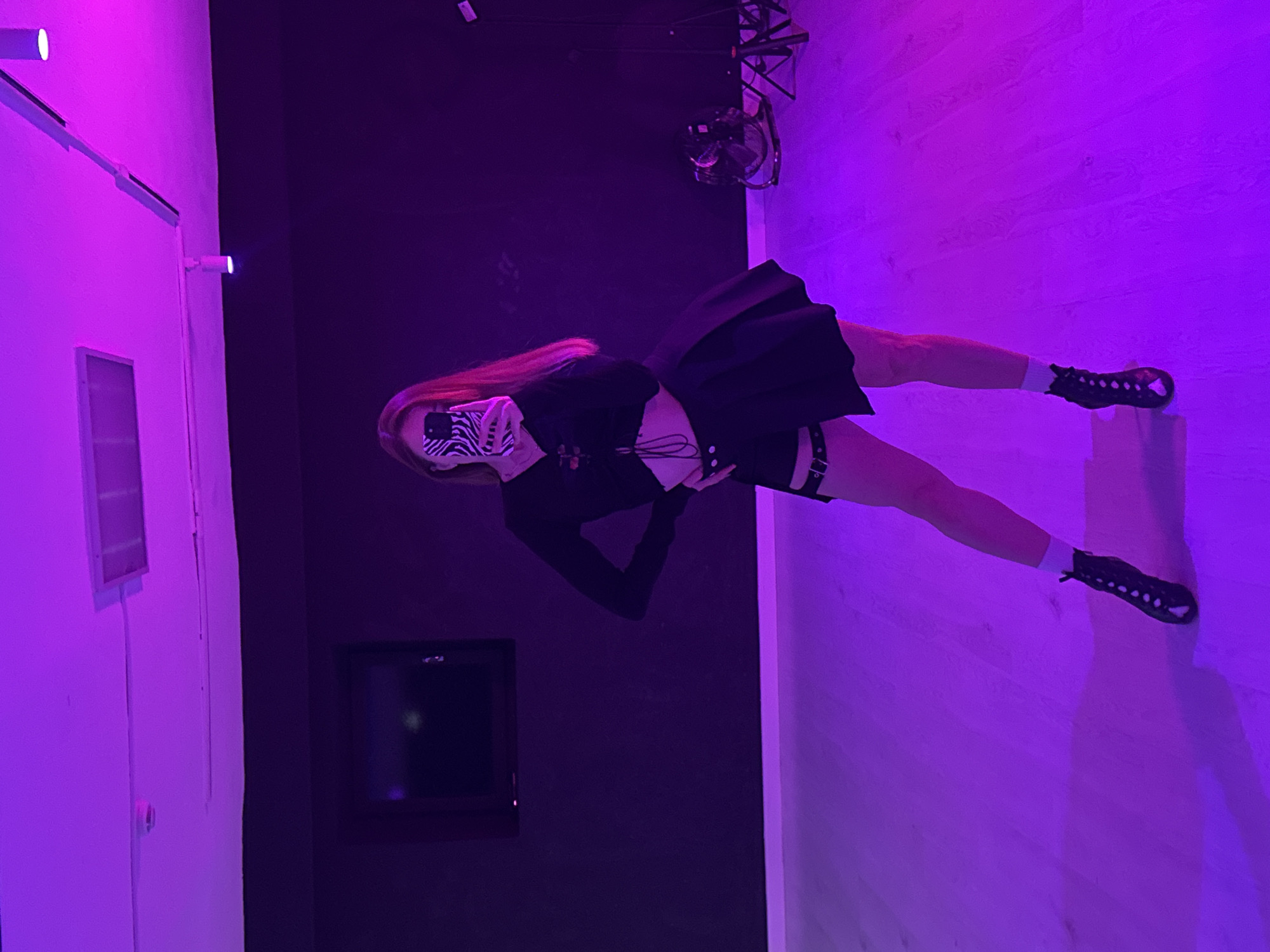 The image captures a person posing in an indoor setting, bathed in vibrant purple and magenta lighting. The main subject is a young individual, standing with their right foot elevated on a white block. They are dressed in a long-sleeved black crop top, a black pleated mini skirt, white socks, and black chunky shoes. Their face is partially obscured by a zebra-print mask, and they are holding a smartphone with a matching zebra-print case up to their face, suggesting they are either taking a selfie or checking the screen. Long reddish-brown hair flows over their shoulders.
The setting includes a light-colored wooden floor and plain walls. One wall, illuminated in bright purple, appears white, while the wall directly behind the person is dark, almost black, and features a mounted rectangular screen or television. The ceiling reveals a fan and some metallic scaffolding. Several light fixtures are visible on the brightly lit walls. The dominant purple illumination creates a dramatic and stylized atmosphere, indicative of an art installation, a photo studio, or an event venue. The pose and attire suggest a fashion-oriented context. No discernible text is visible in the image. The location is Chaikovsky, Russia.