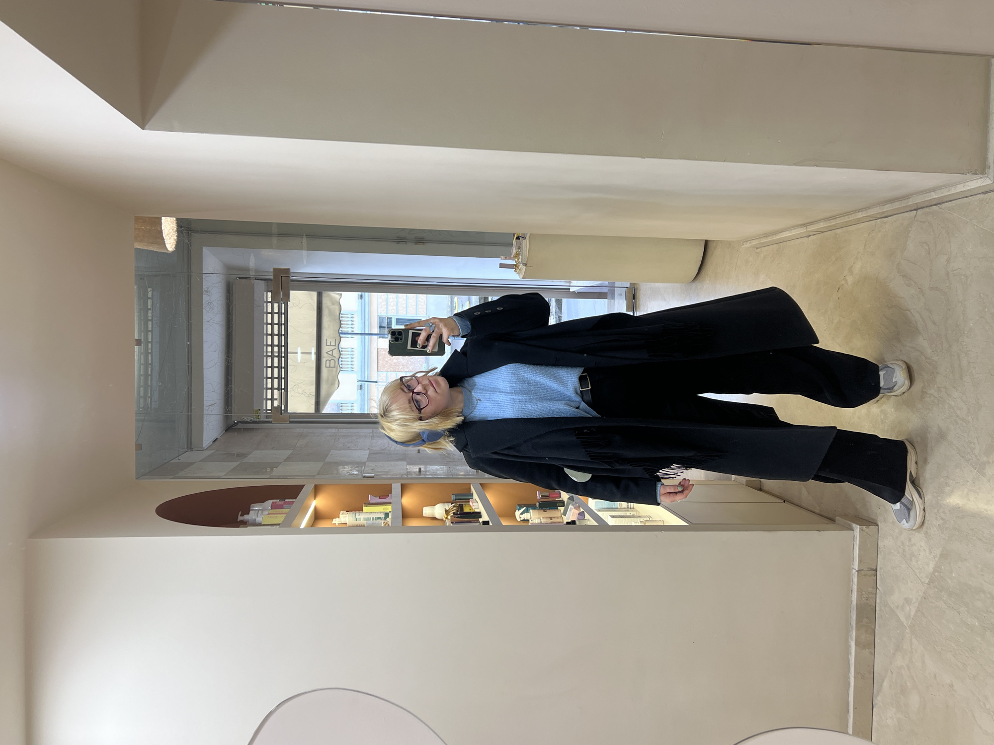 A woman with blond hair, wearing glasses, a blue sweater, black pants, a black coat, and white sneakers, is standing in a room with light beige walls and floor. She is holding a smartphone and taking a picture of herself in a mirror. The mirror reflects a doorway leading to a street scene. Through the doorway, one can see buildings and a partially visible sign with the letters "BAE" on it, suggesting a location in Odesa, Ukraine, due to the presence of Cyrillic characters. There are shelves with products displayed behind the woman.