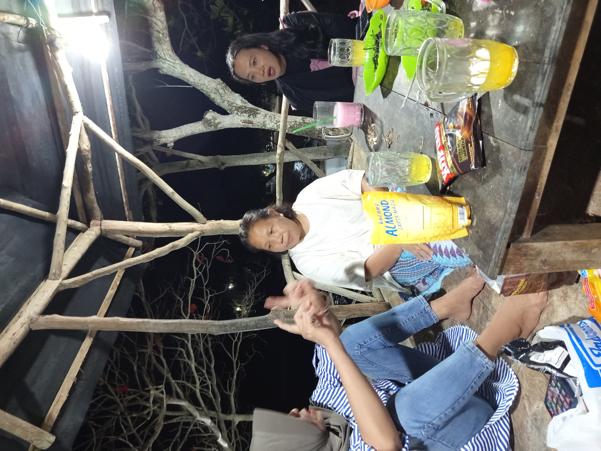 In a dimly lit outdoor pavilion with a wooden roof and support beams, three individuals are gathered around a rustic wooden table. The person on the left, wearing a hijab and a blue and white striped shirt with jeans, is seated with their legs crossed. They are gesturing with their hands, as if speaking. To their right, a woman in a white t-shirt and patterned skirt is seated and looking towards the person on the left. In the background, a third person with long dark hair is seated and looking towards the viewer, wearing a black top.
On the table are several drinking glasses, some containing yellow liquid, and one with a pink beverage. A bag of "Kacang Almond Lapis Madu" (Almond Honey Layered Nuts) and a packet of coffee are also visible. A green plate holds some dark green leafy vegetables or herbs. The environment suggests a casual outdoor dining or social gathering, possibly in a rural or coastal area given the presence of trees with bare branches in the background, which are silhouetted against the dark night sky. The overall impression is one of relaxed interaction among the individuals.