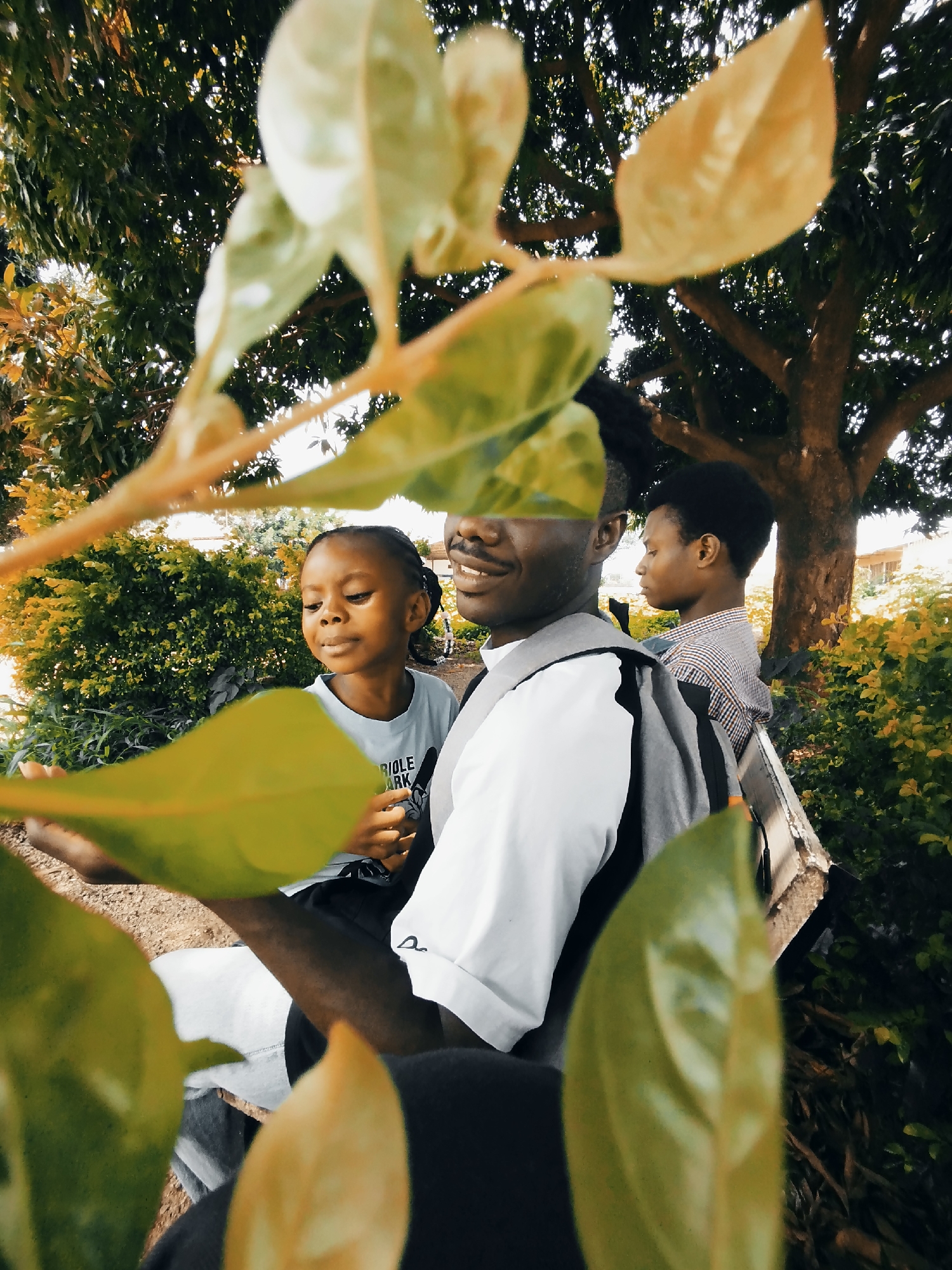 The image shows three young Black individuals outdoors, seated on a bench beneath a large tree. In the foreground, leaves obscure part of the scene. A young girl with braided hair is looking to her left. To her right, a young man with dreadlocks and a backpack smiles broadly, looking towards the girl. Further in the background, another young man with short hair is visible, wearing a plaid shirt and looking to his left. The setting appears to be a park or a garden, with dense green bushes and the trunk of a large tree dominating the background. The lighting suggests it is daytime.
