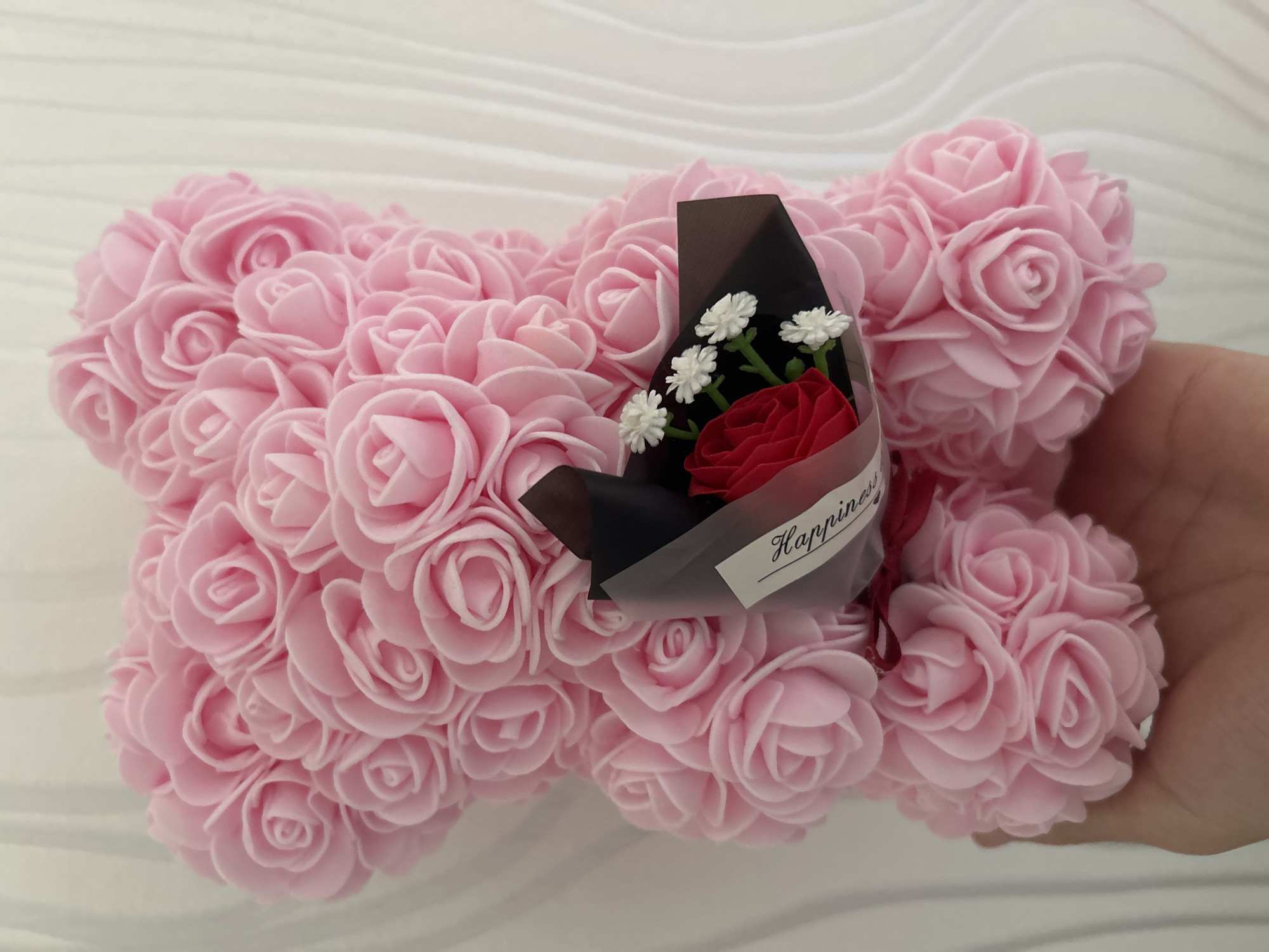 A close-up photograph shows a teddy bear made of pink roses. The teddy bear is being held by a hand on the right side of the image. On the left side of the bear, near its head, is a small bouquet of a single red rose and three white daisies wrapped in black tissue paper. The bouquet is also labeled with a sticker that reads "Happiness" in script. The background is a blurred white surface. The photo was not taken in Strasbourg, France.