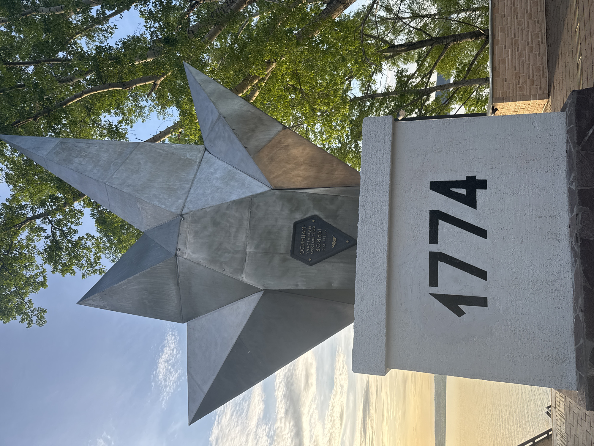 The image depicts a monument in the city of Osa, Russia, commemorating the participants of the Peasant War of 1773-1775. The monument features a geometric star made of metal and a plaque written in Russian. The number "1774" is painted on the side of a white, textured pillar. The background includes trees and a cloudy sky. The monument is located outdoors.