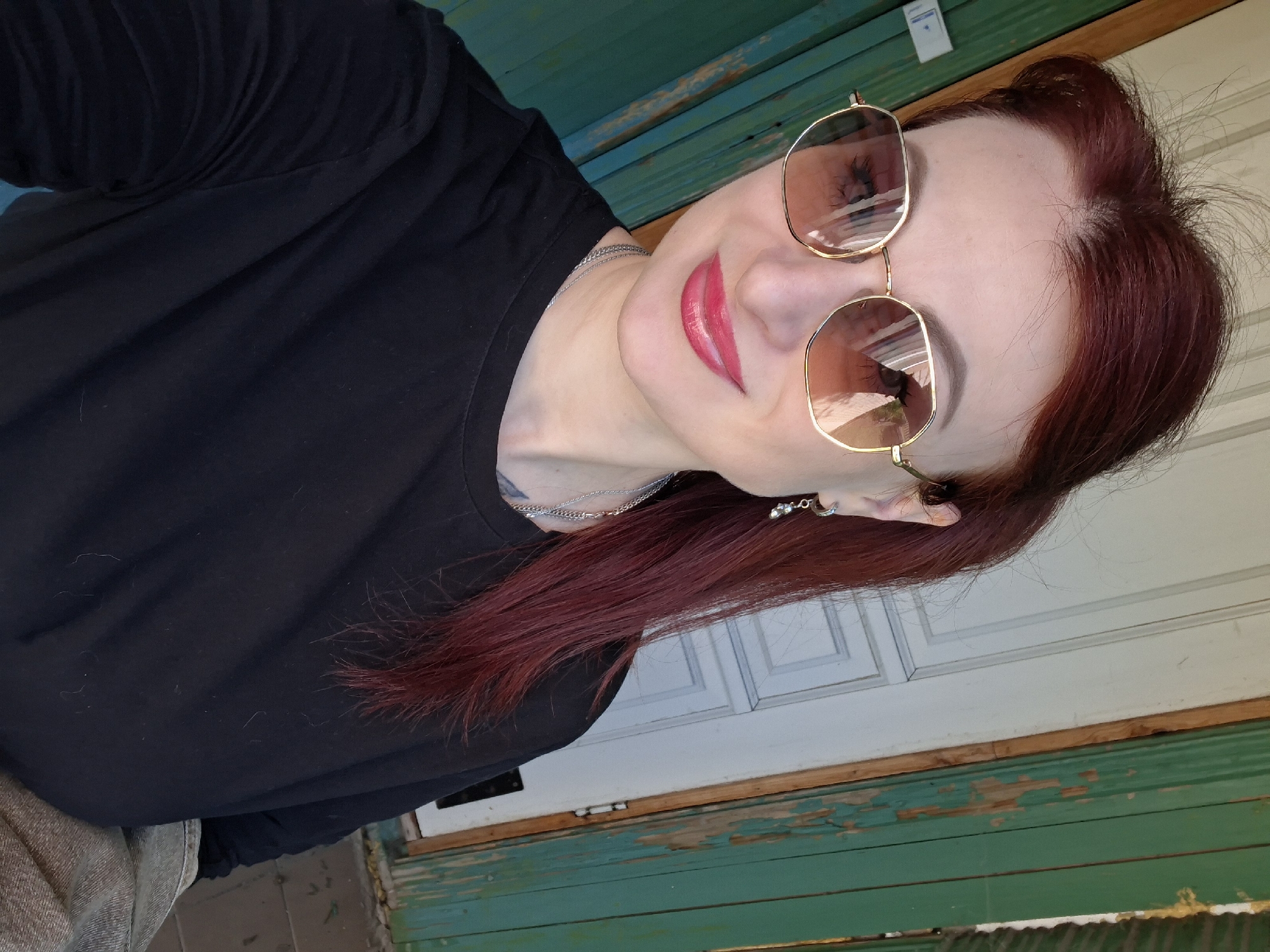 The image shows a woman with red hair, wearing sunglasses and a black top, in Забалоцкі сельскі Савет, Belarus. She is smiling and looking towards the camera. The background appears to be a weathered building exterior with a green and white color scheme.