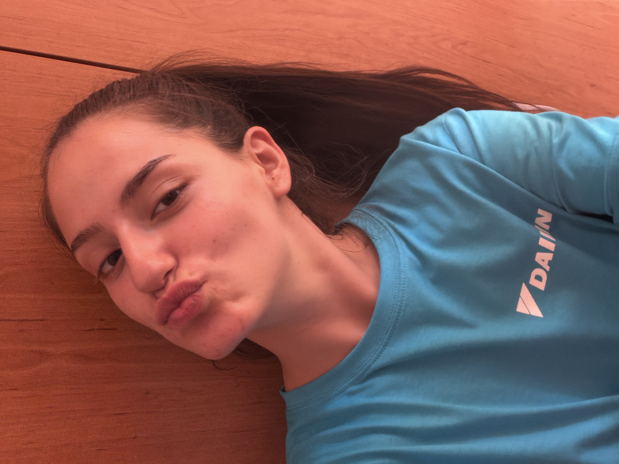 This is a close-up, overhead, selfie-style photograph of a young woman lying on a wooden surface. She has long, dark brown hair pulled back in a ponytail, and is wearing a bright blue t-shirt with a pink logo that reads "DAIKIN" on the chest. Her face is tilted towards the camera, with her lips pursed in a playful "kiss" expression. Her eyes are looking slightly away from the camera. The lighting suggests it could be daytime, and the warm tone of the wooden surface implies an indoor setting. The woman's expression and pose convey a lighthearted and casual mood. The wooden surface appears to be a floor or tabletop.