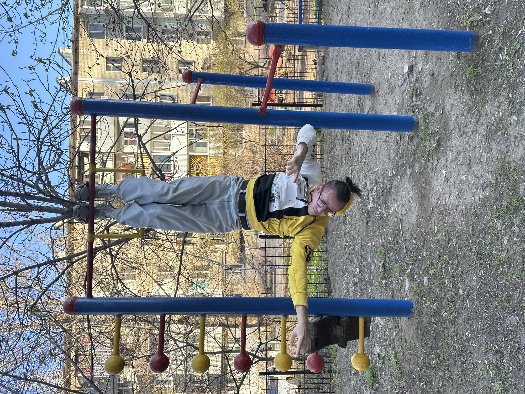This image captures a person hanging upside down from blue horizontal bars at an outdoor playground, likely performing an exercise. The individual, appearing to be a young adult, is wearing a yellow and white hoodie, grey pants, and light-colored sneakers. Their feet are hooked over an upper bar, while their right hand reaches towards a lower bar featuring alternating red and yellow ball-shaped hand grips. Their left hand is raised, making a gesture with fingers. The person wears glasses, and their hair is tied back.
The scene is set in an outdoor recreational area. In the background, leafless trees with visible new leaf buds suggest it is springtime. Behind the trees, several multi-story residential buildings are visible, indicating an urban or suburban setting. The ground beneath the playground equipment is dirt and gravel, with patches of sparse grass. The sky is clear and blue, typical of a sunny daytime. The overall impression is one of physical activity in a public park or courtyard, consistent with locations in Saratov, Russia. Text is visible on the person's clothing but is not clearly legible.