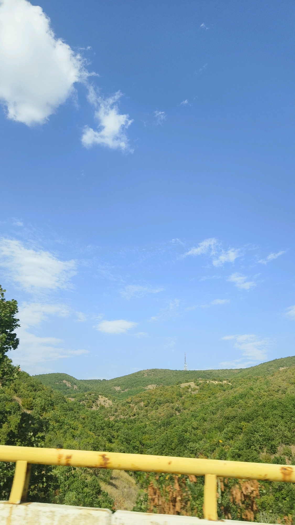 The image depicts a view of rolling green hills covered in dense forest under a bright blue sky with scattered white clouds. In the foreground, a yellow railing, likely part of a bridge or overpass, runs horizontally across the lower portion of the frame. The railing shows signs of rust and wear. To the left, a portion of a large tree with green foliage is visible. In the distance, atop one of the hills, a tall, thin tower, possibly a telecommunications tower, is silhouetted against the sky. The scene suggests a natural landscape, possibly on the outskirts of a town or city. This view is consistent with the geography of Letevci, North Macedonia, known for its hilly terrain and vegetation.