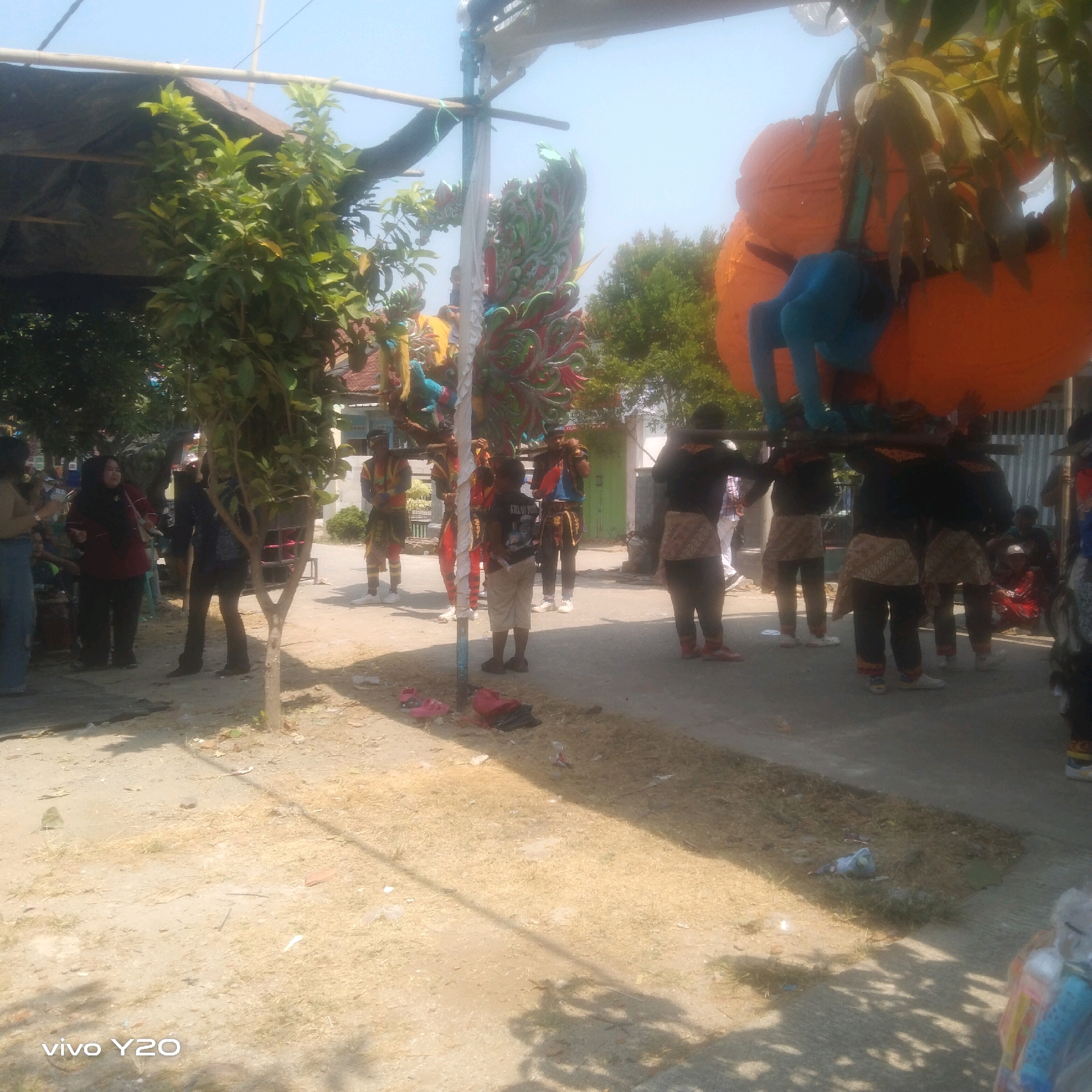 A group of people are gathered outdoors, possibly for a festival or parade, in an urban setting. In the foreground, a sandy area with sparse dry grass is visible, with shadows of trees cast across it. A small tree with green leaves stands in the middle ground. Further back, a paved area shows several individuals dressed in traditional attire, some carrying large, colorful decorations. One prominent decoration is a large orange, spherical object supported by poles. In the background, there are buildings, trees, and a clear sky. Several people are observing the scene, while others are actively participating in carrying the decorations. The overall impression is of a community event taking place on a bright, sunny day.