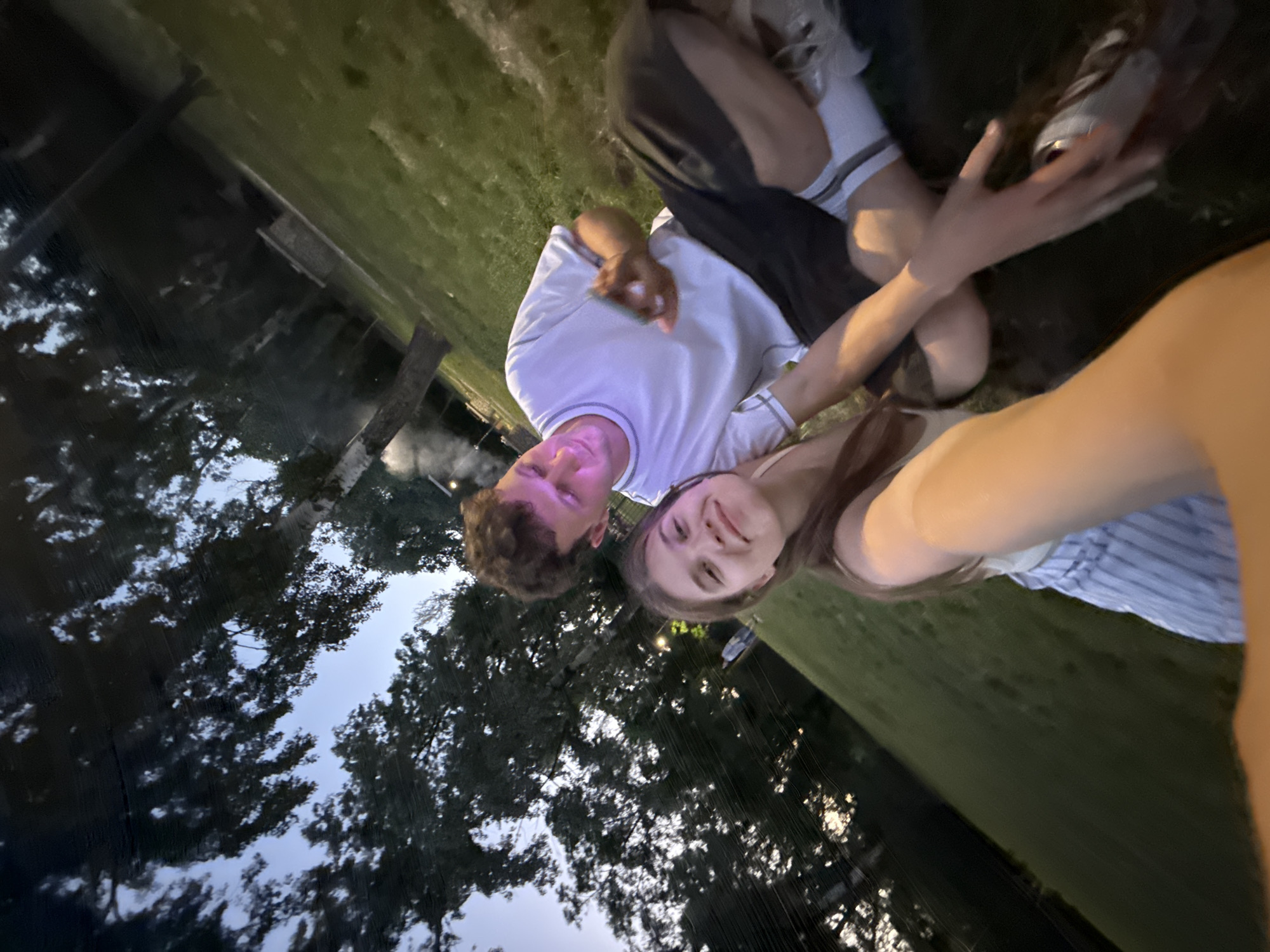 A photograph taken in Cukarica Urban Municipality, Serbia, at twilight shows a young man and woman reclining in a grassy area near trees. The man is wearing a white shirt and the woman is wearing a striped top. The photo is taken from a low angle and rotated counterclockwise.