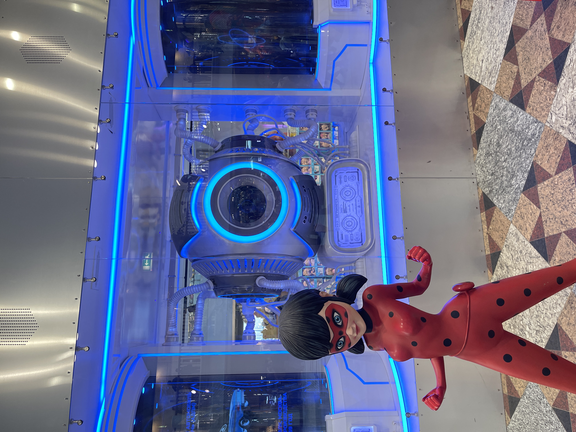 The image depicts a statue of Ladybug, a character from the animated series "Miraculous: Tales of Ladybug & Cat Noir," standing in front of a futuristic-looking machine encased in glass. The machine has a large lens-like structure with blue neon lighting around it. The statue is red with black spots, mirroring Ladybug's superhero costume. The background includes a metallic wall on the left and a patterned floor on the right. The location is Moscow, Russia, as indicated by Russian text visible within the glass enclosure.