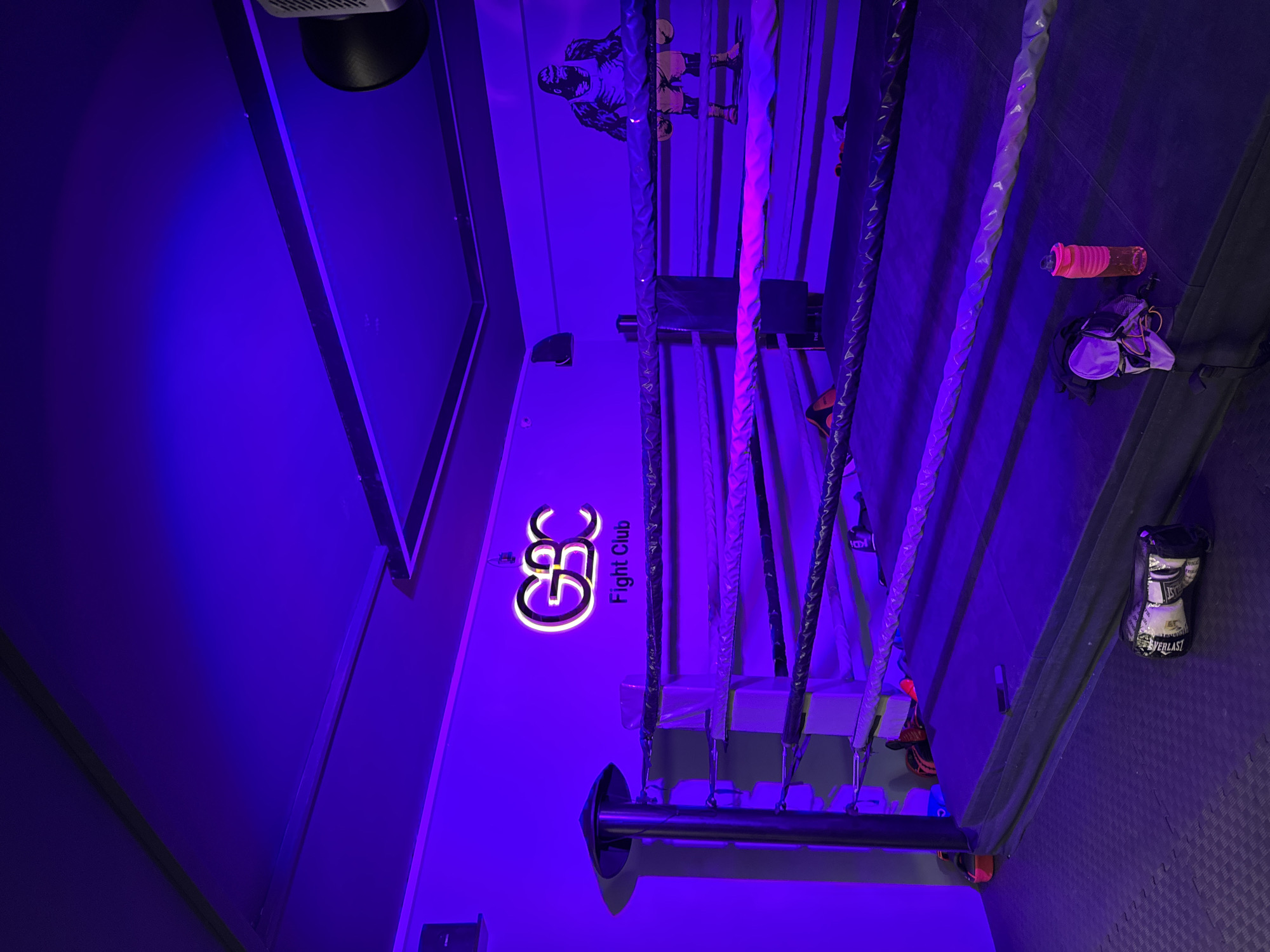 This media file depicts a boxing ring inside a dimly lit, neon-lit gym. The scene is bathed in purple and pink hues from the ambient lighting, creating an intense atmosphere. The camera is angled from the top of the ring looking down, with the ropes of the ring prominently featured in the foreground.
Visible objects include boxing gloves, a water bottle, and possibly some training equipment. On the wall behind the ring, there is a large stylized logo that reads "GBC Fight Club". A graphic of a boxer is also visible on the wall. The overall setting suggests a dedicated space for boxing training or competition.
There are no people visibly present in the frame, but the presence of the equipment and the branded signage implies that the location is actively used for its intended purpose. The time of day is indeterminate due to the artificial lighting, and no weather cues are present. The scene evokes a sense of determination and grit associated with the sport of boxing. The location context is Kâğıthane, Turkey.
