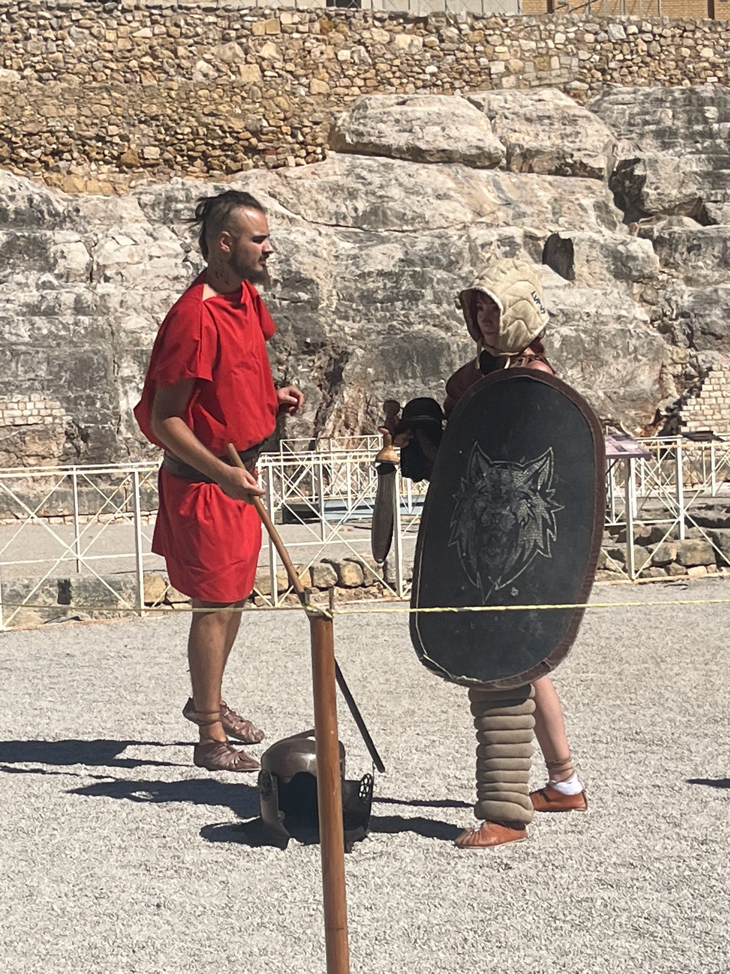 Two individuals, dressed in what appear to be Roman gladiator costumes, stand on a gravel surface in front of a rocky, stone-built amphitheater. The amphitheater's tiered seating and stone construction suggest a historical site. The person on the left wears a red tunic and leather sandals, holding a wooden staff. The person on the right wears a padded helmet, a protective greave on their right leg, and holds a large, oval shield adorned with a wolf's head emblem. A gladius, or short sword, is visible at their hip. The ground is covered in a fine, light-colored gravel, with shadows cast by the individuals and other unseen objects. A white barrier with diamond-shaped openings runs horizontally across the background, separating the performance area from the wider environment. The overall scene depicts a re-enactment or demonstration related to Roman history, likely taking place at the Amphitheatre of Tarraco, a UNESCO World Heritage site in Tarragona, Spain.