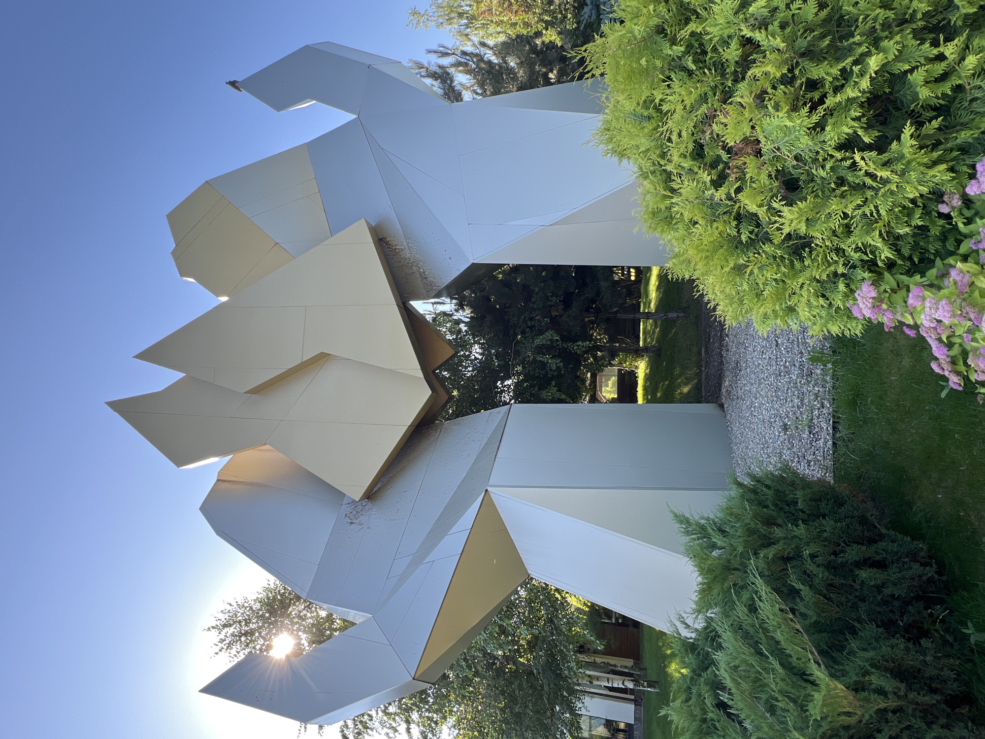 A large, abstract sculpture made of metallic, angular panels stands outdoors. The sculpture is primarily silver and gold, with some darker areas that may indicate weathering or shadow. It has a complex, geometric design that resembles large, interlocking hands or abstract wings reaching upwards. The sculpture is situated in a park-like setting with green grass, lush bushes in the foreground, and trees in the background. The sun is shining brightly, casting lens flare through the sculpture and illuminating the scene. A bird is perched on the upper portion of the sculpture. The ground beneath the sculpture appears to be covered in gravel. The overall impression is of a modern art installation in a natural environment. This sculpture is located in Plastunivska, Russia.