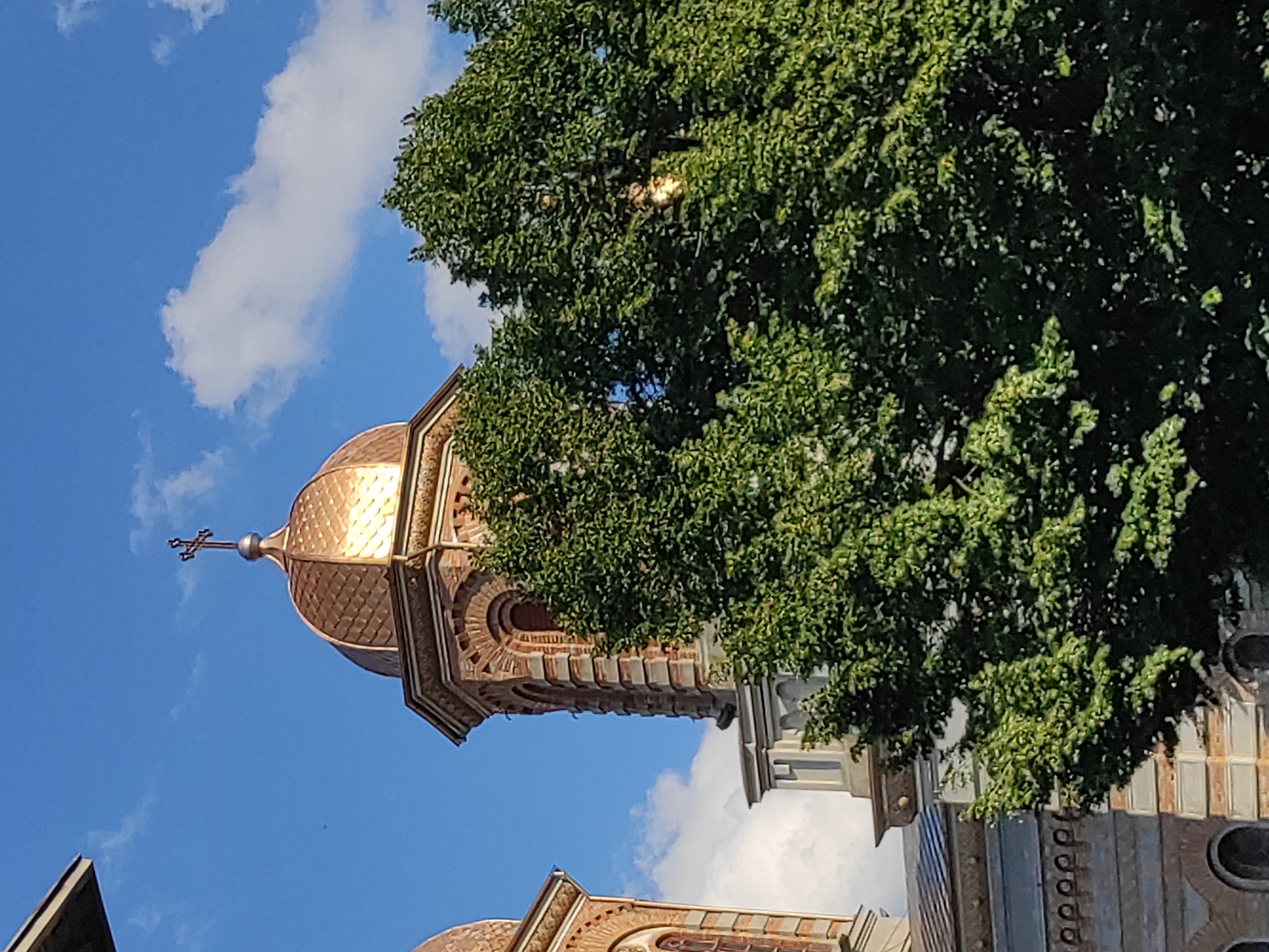 The image captures the dome of St. Andrew's Cathedral in Constanța, Romania, on a clear day. The copper dome, adorned with a cross, reflects the sunlight, contrasting with the blue sky and scattered white clouds. A large, lush green tree partially obscures the view of the cathedral's tower and facade. The architectural style of the cathedral is visible, with brickwork and arched windows. The scene is tranquil, with no people or activity evident.