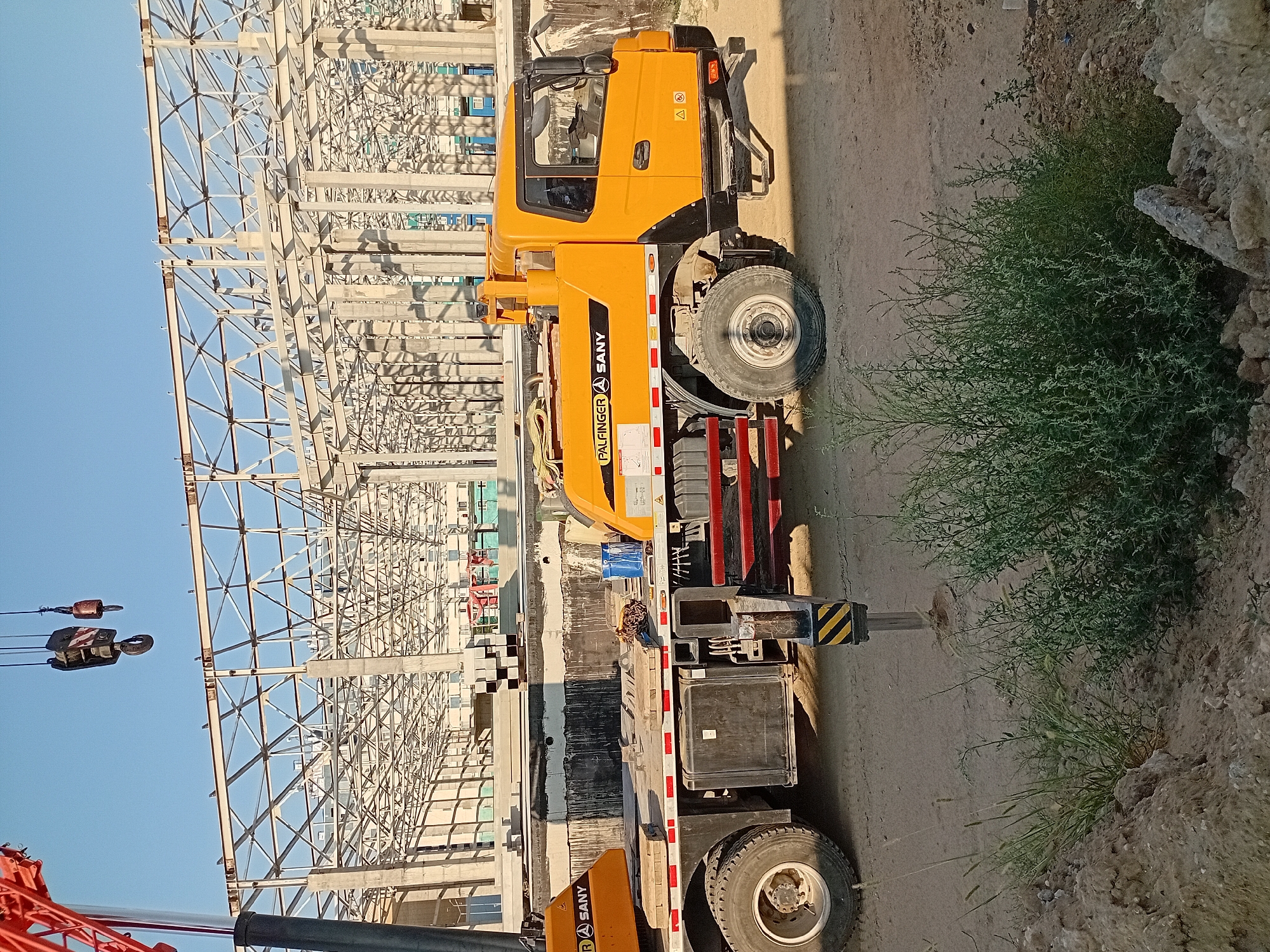 An orange crane truck, labeled "PALFINGER SANY," is parked on a dirt construction site. The truck is facing right and has its outriggers deployed. Behind the truck, a large steel structure, presumably part of a building under construction, is visible against a clear blue sky. In the foreground, there are piles of dirt and some green vegetation. The location is identified as the city of Semey, Kazakhstan, indicating this is an outdoor construction scene.
