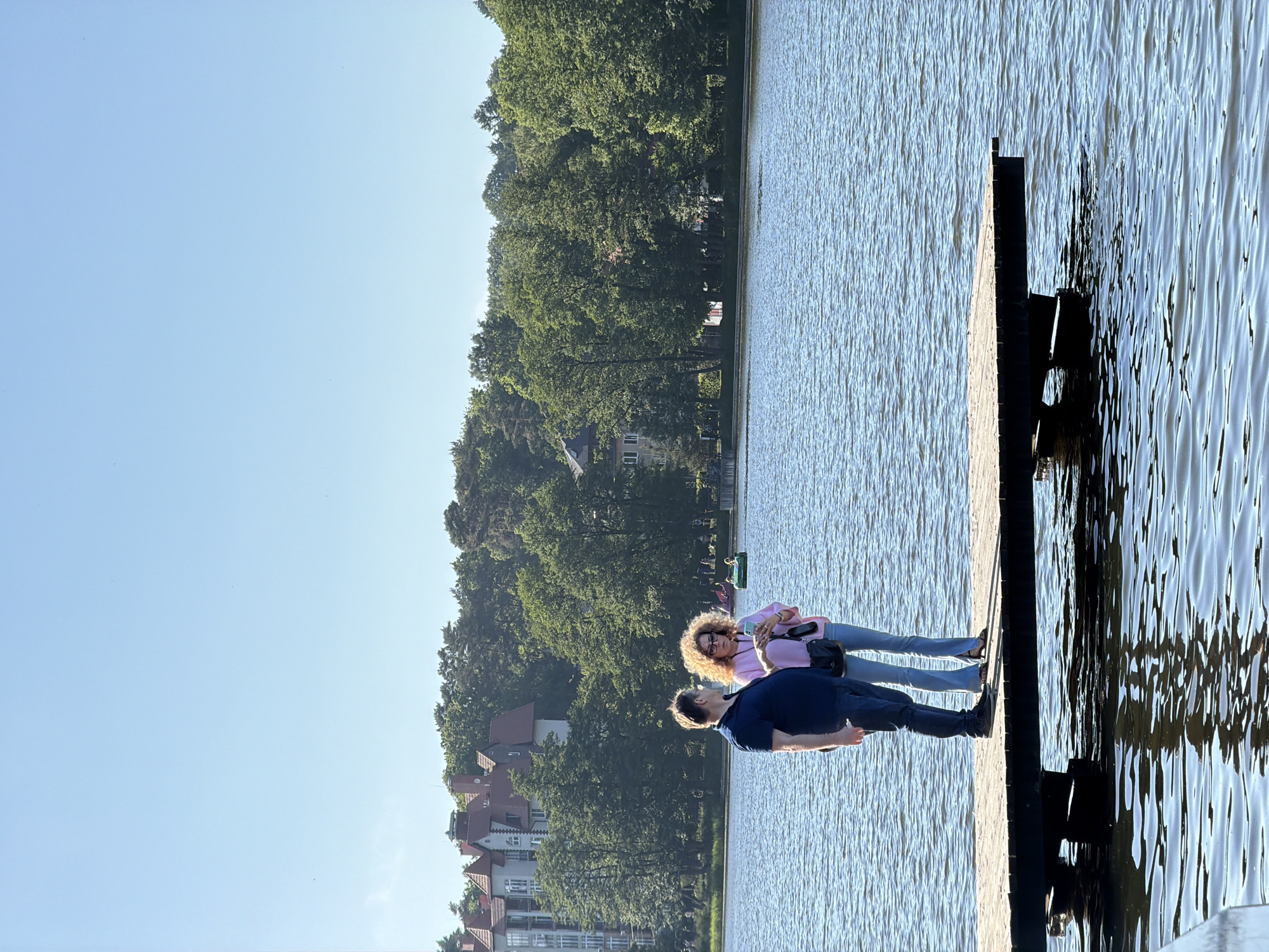 This media file depicts two people standing on a wooden pier by a body of water. The person in the foreground on the right is wearing a dark blue shirt and dark pants, with their head turned upwards to look at the person on the left. The person on the left, also facing towards the right, is wearing a pink jacket and light blue jeans, with blonde curly hair and glasses. They appear to be holding a phone.
The scene is set with a clear blue sky above, and a shoreline lined with lush green trees in the background. Several buildings with red roofs are visible amongst the trees, suggesting a populated area. The water is calm with ripples, reflecting the sunlight. The overall impression is of a bright, sunny day, likely in the late afternoon or early evening given the angle of the sun and the warm lighting. The atmosphere seems relaxed and pleasant.
The orientation of the image is landscape, but it has been captured as if the camera was rotated 90 degrees clockwise. The location is identified as Svetlogorsk, Russia. There is no visible text in the image.