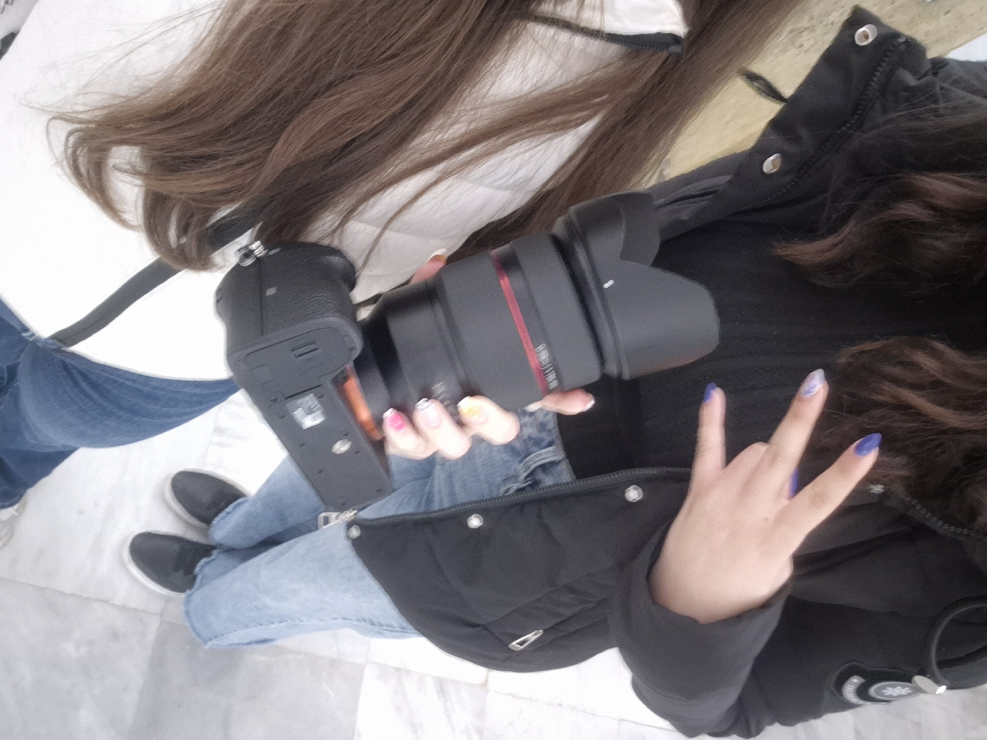 A first-person perspective photograph shows two people indoors on a white tiled floor. The person taking the photo is wearing blue jeans and black slip-on shoes, and holding a black digital camera with a large lens in their left hand. Their nails are painted with various colors. They are also wearing a black puffer jacket over a black shirt. The other person is wearing blue jeans and a white top. Both people have brown hair. The location is likely indoors, as the floor is tiled. Given the request for a location in Srebarna, Bulgaria, and the absence of external environmental cues, this photo could be taken in a house, hotel, or other indoor facility in Srebarna.