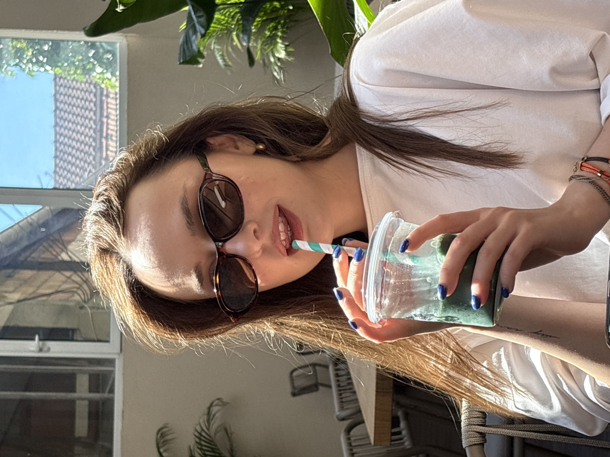 In this medium shot photograph, a young woman with long, auburn hair is seated outdoors, holding a clear plastic cup with a green beverage and a blue and white striped straw. She is wearing sunglasses with dark, oval frames and a white t-shirt. Her nails are painted a vibrant blue. Her left hand is wrapped around the cup, while the straw is positioned between her lips. The background suggests an outdoor seating area, possibly a cafe, with glimpses of furniture, plants, and windows reflecting a sunny sky and rooftops. The lighting indicates it is daytime, with strong sunlight casting shadows and highlighting the subject. The overall scene conveys a relaxed, casual moment. There is no specific indication of the location being in the city of Kragujevac, Serbia.