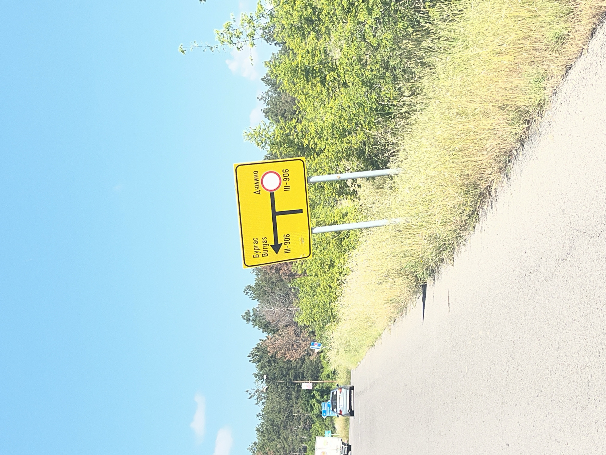 The image shows a yellow directional sign in Tankovo, Bulgaria, pointing to two different locations. On the left side, the sign indicates "Бургас" (Burgas) and "Burgas" with an arrow pointing left, followed by "III-906". On the right side, it indicates "ДЮЛИНО" (Dyulino) with a road junction symbol and a circular sign with a red border, followed by "III-906". The sign is supported by two metal poles. The surrounding environment consists of a paved road, dry grass, green bushes, and a dense forest of coniferous trees in the background under a clear blue sky. A silver car is visible on the road to the left, and a white truck is further in the distance.
