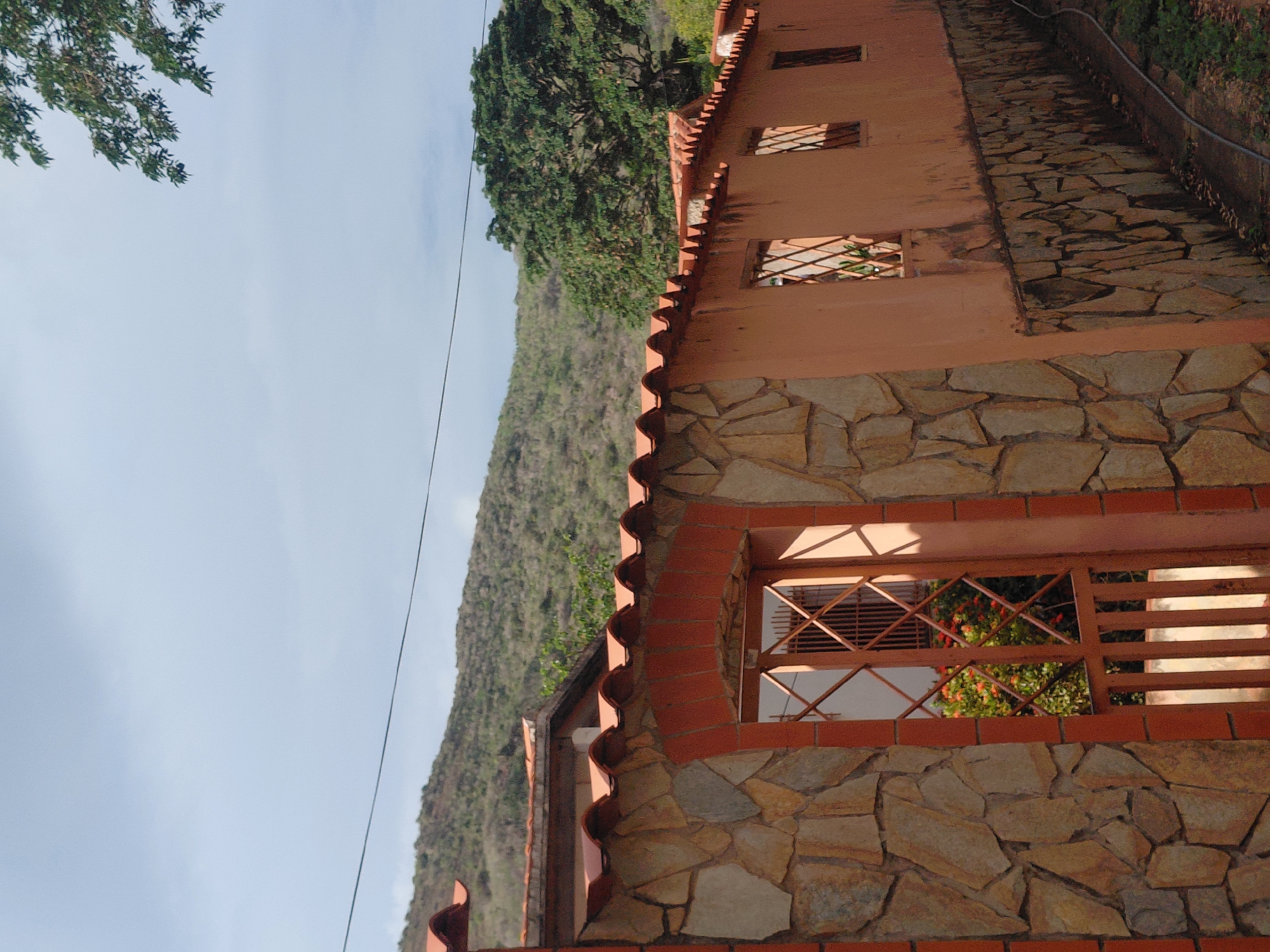 The image depicts an exterior view of a property wall in Santa Ana, Venezuela. The wall is constructed from a combination of rough-hewn stones and a peach-colored stucco. It features an arched entryway with a decorative metal gate, which reveals a glimpse of lush greenery and red flowers behind it. Several small, barred windows are visible along the length of the wall. The top of the wall is capped with terracotta roof tiles. In the background, a gently sloping, tree-covered hill rises against a pale, overcast sky. The ground along the base of the wall is a mixture of dirt and scattered leaves, with a black cable or hose running along it. The overall impression is one of a rustic and secluded residential or rural setting.