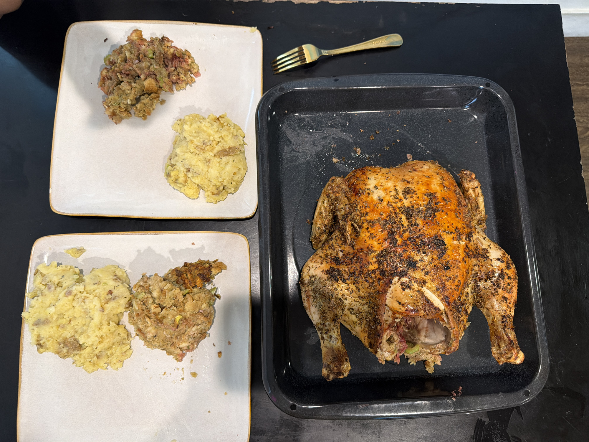 The image shows a cooked chicken in a black baking pan, a gold fork, and two plates with mashed potatoes and stuffing. The plates are white with a gold trim. The food is arranged on a black surface. The location is likely a home kitchen in Los Angeles, United States of America.