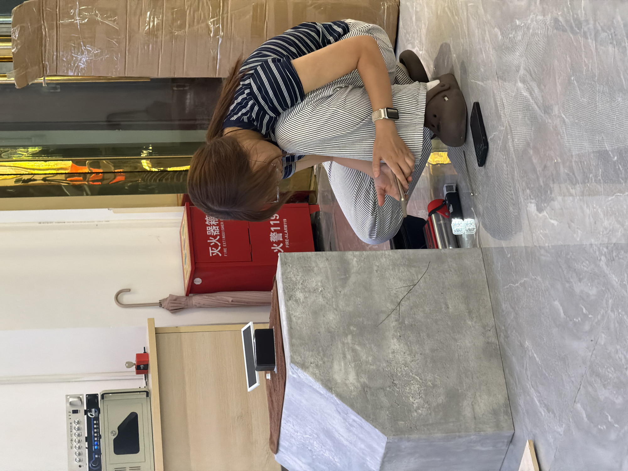 A person is crouching on a gray tiled floor in what appears to be an indoor space, possibly a shop or workshop, in Jiangjin, China. The individual, wearing a striped t-shirt and striped pants, is looking down at a smartphone they are holding. To the left of the person is a large, angled concrete structure. Behind the concrete structure, there is a red fire extinguisher cabinet with Chinese characters and an umbrella leaning against it. Further to the left, on a wooden counter, are various electronic devices and a small red item. The background features a reflective surface, likely a window or door, showing some yellow and orange light reflections, and a large cardboard box wrapped in plastic tape.