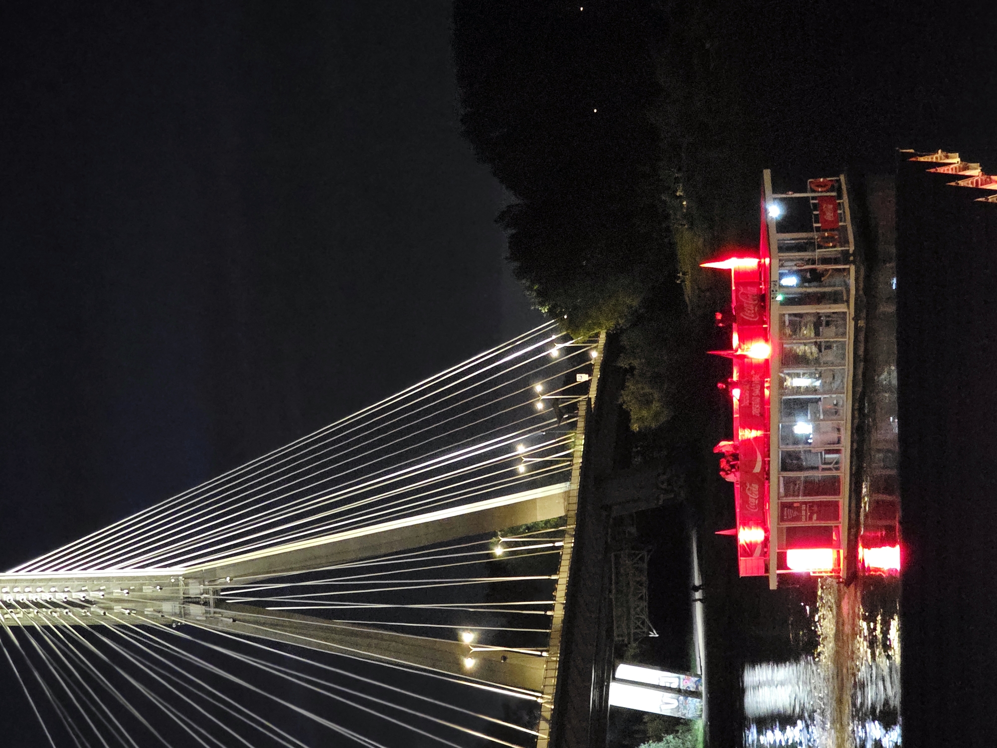 A night scene in Warsaw, Poland, features the Świętokrzyski Bridge illuminated by bright lights. The bridge's cables are stark white against the dark sky, and lights line the bridge's walkway. Below the main bridge structure, another bridge supports a road, with graffiti visible on its concrete supports.
In the foreground, a red-lit barge, likely a floating restaurant or bar, travels on the Vistula River. The barge has large windows and is adorned with Coca-Cola branding. Several people are visible on the upper deck of the barge. The water reflects the barge's red lights, creating a glowing trail. Trees and dark foliage line the riverbank in the background.