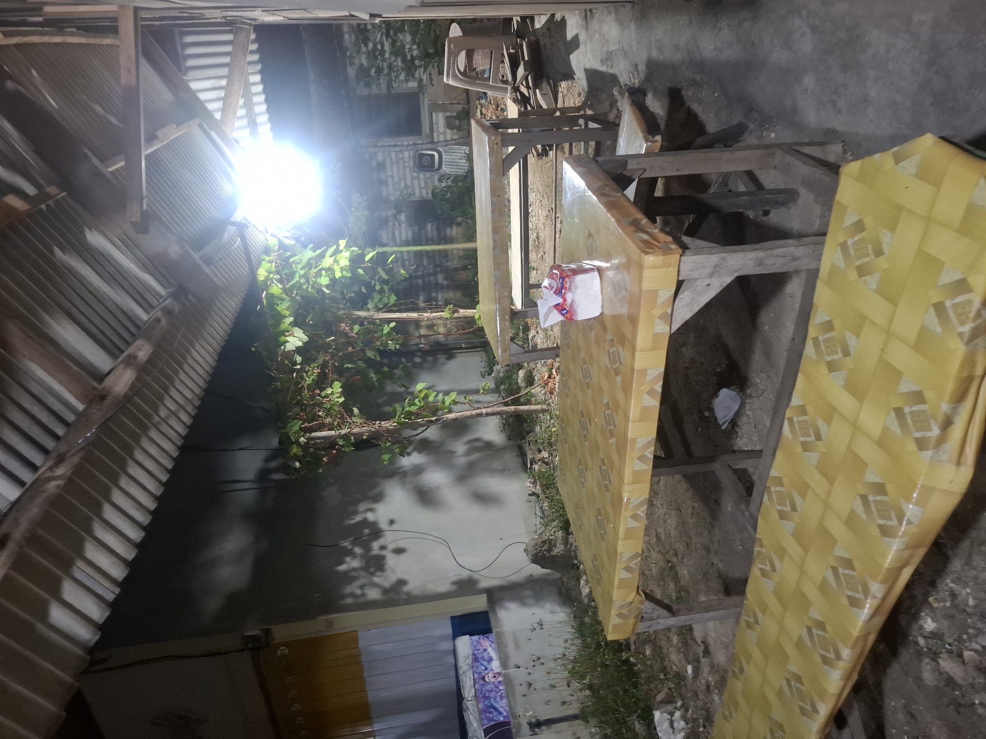 The image depicts an outdoor seating area at night, likely at a roadside establishment or warung in Bulubrangsi, Indonesia. Two wooden tables are visible, each covered with a yellow tablecloth featuring a repeating geometric pattern. The table in the foreground has a box of tissues on it. In front of the foreground table, a matching yellow bench is partially visible. In the background, another table with benches is set up. A bright overhead light illuminates the scene, casting shadows from the trees and the corrugated metal roof overhead. The environment appears somewhat unkempt, with dirt ground and scattered debris. To the left, a building wall is visible with some decorative elements and what appears to be a large framed image. In the background, there are more structures, including what looks like discarded furniture or building materials, and a ventilation unit on a brick wall. No people are present in the image.
