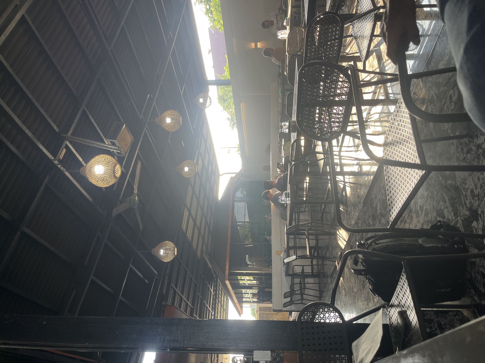 The image depicts an outdoor seating area of a cafe or restaurant, possibly in Glameunasah Baro, Indonesia, with a roof providing partial shelter. The setting is characterized by a blend of natural light and artificial lighting from decorative woven pendant lamps. Sunlight streams in from the right, casting long shadows across the patterned tiled floor and reflecting off the metallic surfaces of chairs and tables.
Several tables with chairs are arranged throughout the space, some occupied by patrons. In the foreground, a dark backpack rests on the floor beside a chair. In the mid-ground, patrons are seated at tables, some engaging in conversation, while others appear to be looking at their phones. One individual is visible standing in the background near an enclosed structure. The architecture suggests a contemporary design with a dark-colored roof and a building with glass elements. The overall ambiance appears relaxed and casual, typical of a daytime gathering spot.