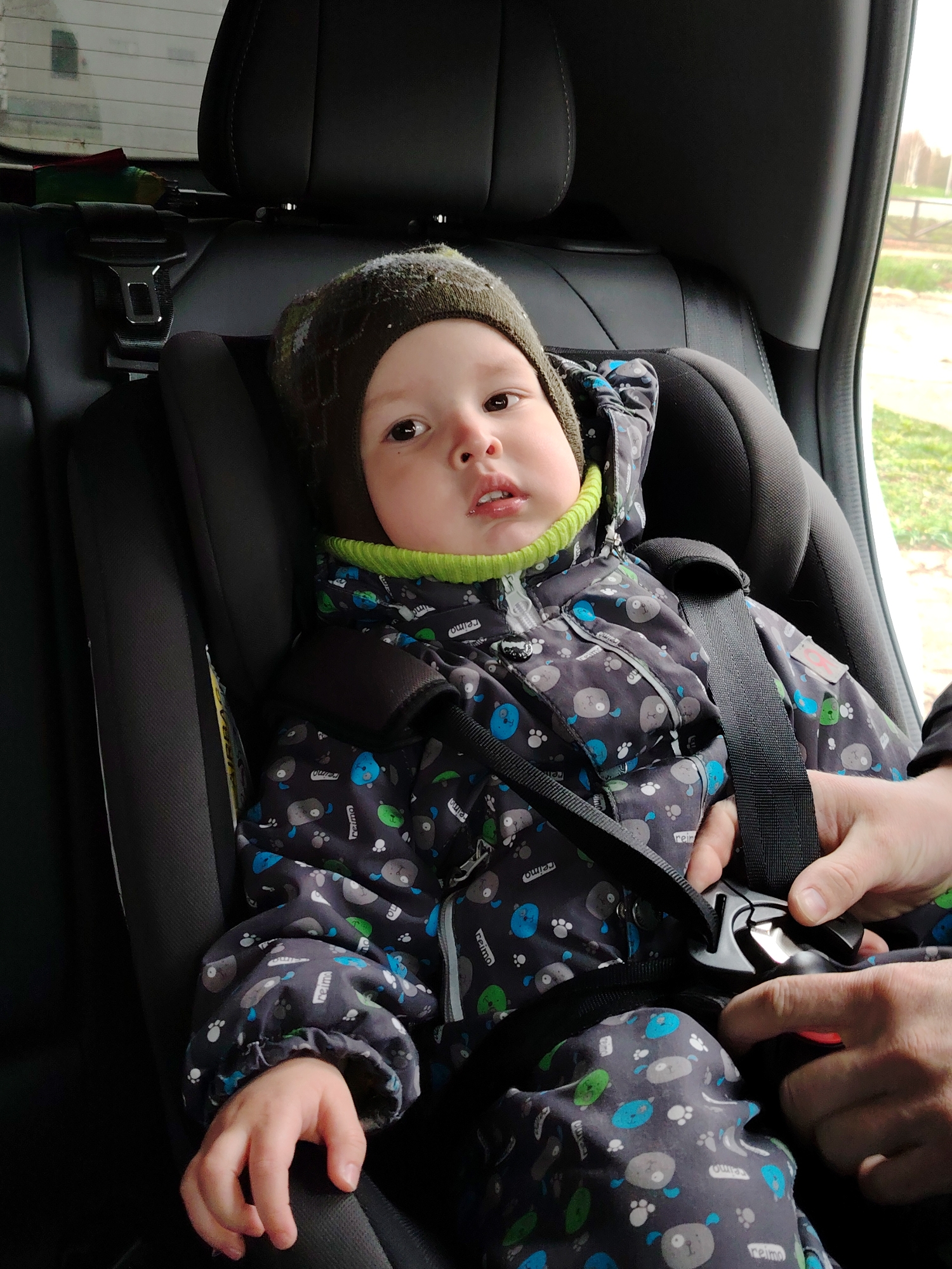 A young child, likely a toddler, is seated in a black car seat in the back of a vehicle. The child is dressed in a warm, dark grey winter jacket adorned with a repeating pattern of blue, grey, and green cartoonish animal figures and paw prints. A bright green neck collar or gaiter peeks out from under the jacket, and a dark green knit hat covers the child's head. The child looks directly at the camera with a neutral, somewhat curious expression.
An adult's hands are visible in the lower right of the frame, actively securing the child's harness in the car seat. They are manipulating the black car seatbelt buckle, which features silver and red accents. The car seat itself is black and appears well-padded.
Through the window on the right side of the car, blurred outdoor scenery is visible, including a light sky and hints of green vegetation, possibly suggesting a rural or suburban setting during daytime. The car's interior features black leather or synthetic upholstery on the seatbacks and headrests. A yellow label with "AIRBAG" printed on it is partially visible on the side of the car seat, and the brand name "reimo" is repeatedly printed on the child's jacket. The scene is located in what is likely Markovskiy, Russia.