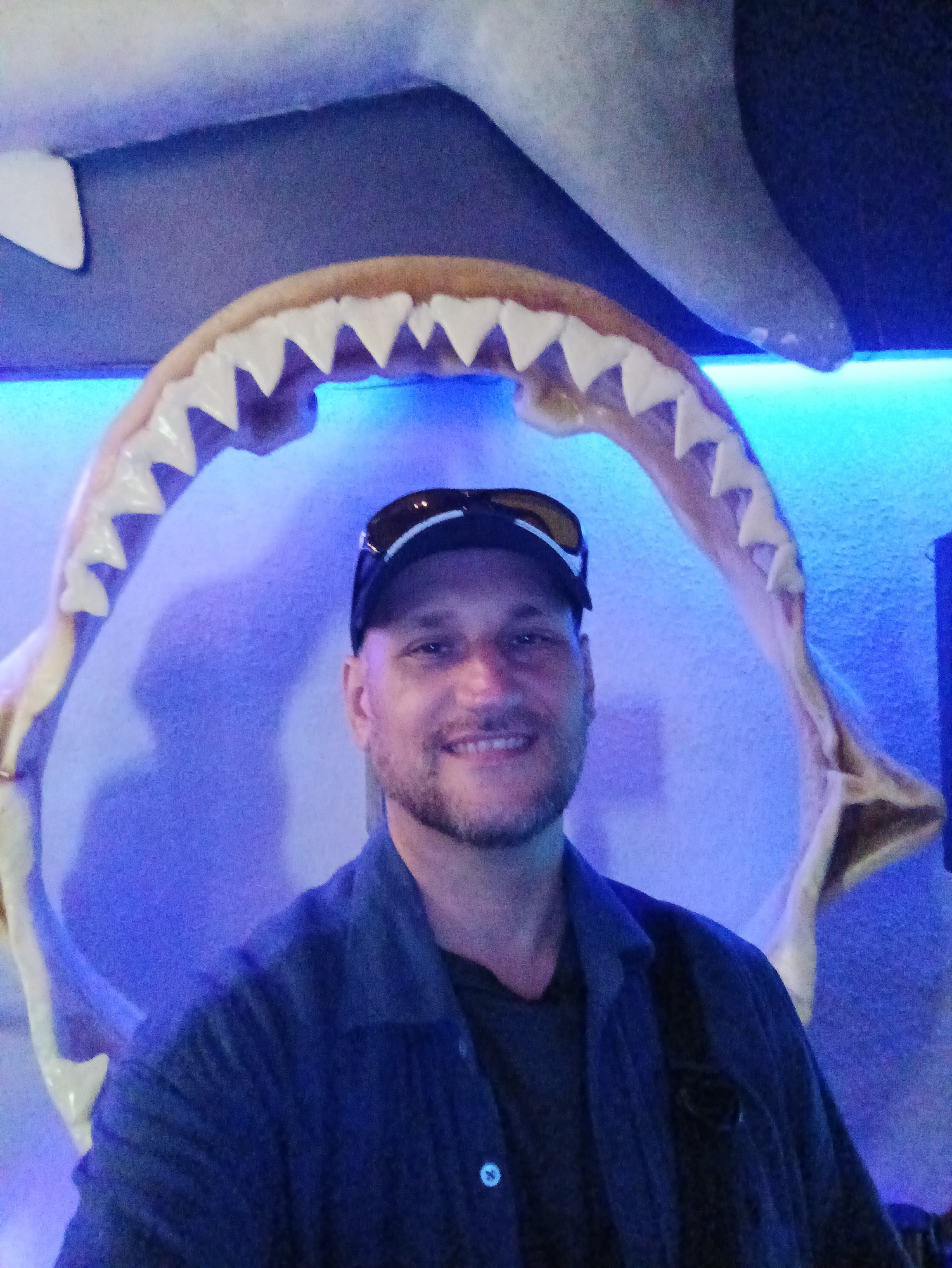 A man with a beard and sunglasses on a baseball cap is posing for a photo. He is smiling and looking at the camera. He is wearing a dark blue collared shirt over a dark grey t-shirt. Behind him, a large replica of shark jaws is displayed. The backdrop is a blue-lit wall. It is not possible to determine the location within Kuala Lumpur, Malaysia based on the provided image.