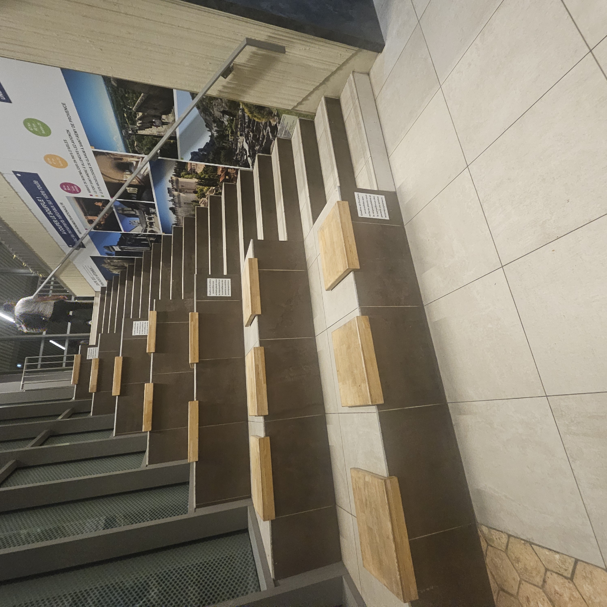 The image depicts an indoor amphitheater-style seating area with tiered steps made of dark gray material. Several wooden planks are placed on some of the steps, serving as seats. A metal railing runs along the right side of the stairs. On the wall behind the seating, a large mural showcases scenic views of Provence, France, featuring historical buildings and landscapes. One sign on the wall reads "A COUPER LE SOUFFLE !" which translates to "Breathtaking!". Another smaller sign on one of the steps displays a warning in French regarding supervision of children. A person wearing a white t-shirt and dark pants, carrying a backpack, is walking up the stairs in the background. The floor in the foreground is tiled with large, light-colored square tiles. The overall environment appears to be a public space, possibly a museum, exhibition hall, or visitor center, located in Lançon-Provence, France, as suggested by the mural's theme.