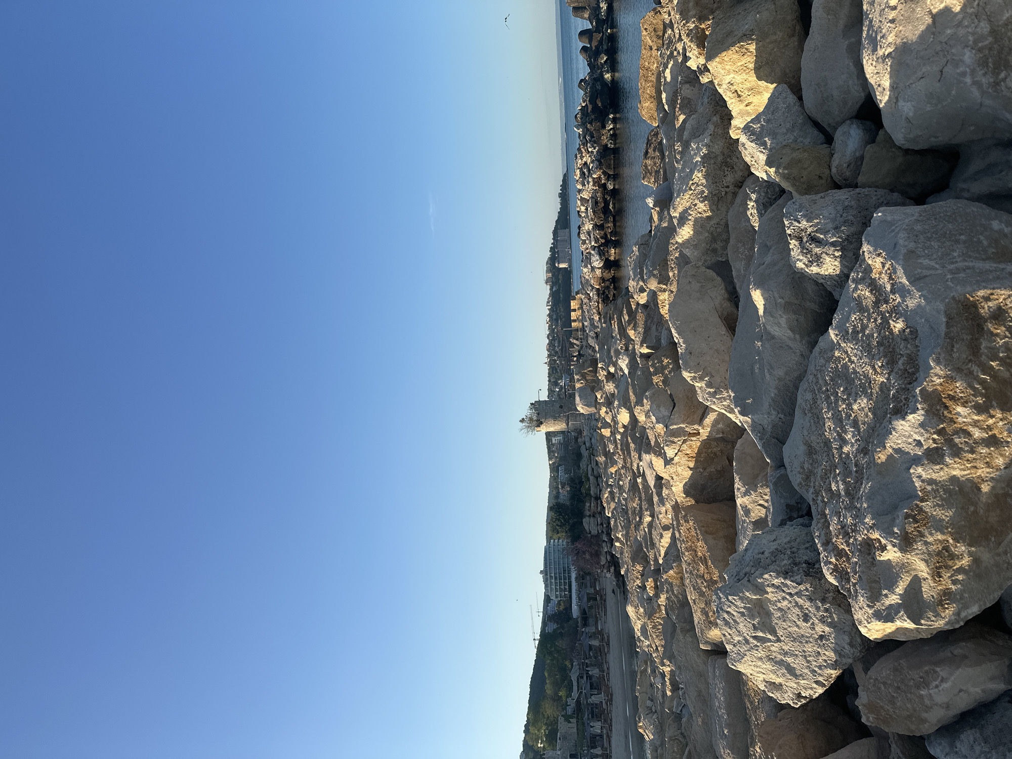 The image depicts a scene in Varna, Bulgaria, featuring a breakwater composed of large, irregular rocks. The rocks are illuminated by the sun, creating shadows and highlighting their textures. In the background, the cityscape of Varna is visible, including buildings, a lighthouse, and a coastline. The sky is clear and blue, with a few faint clouds and a bird visible. The photo is taken at an angle, emphasizing the height of the breakwater and the distance to the city.
