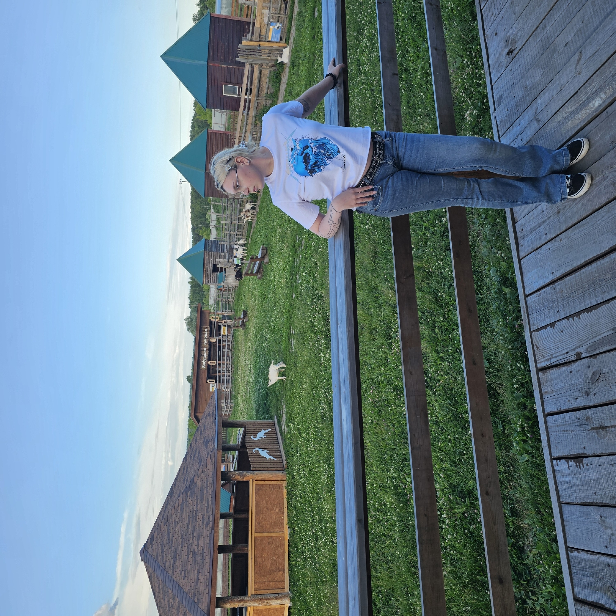 A woman with blonde hair and glasses is standing on a wooden deck, leaning on a railing. She is wearing a white t-shirt with a blue graphic and blue jeans. In the background, there is a grassy field with several small wooden buildings with teal roofs. A white goat is visible in the field. The sky is clear and blue, suggesting it is daytime with fair weather. The scene appears to be a rural or farm setting. Visible text on a building in the background reads "Бабушкин сеновал" (Babushkin senoval), which translates to "Grandmother's hayloft."