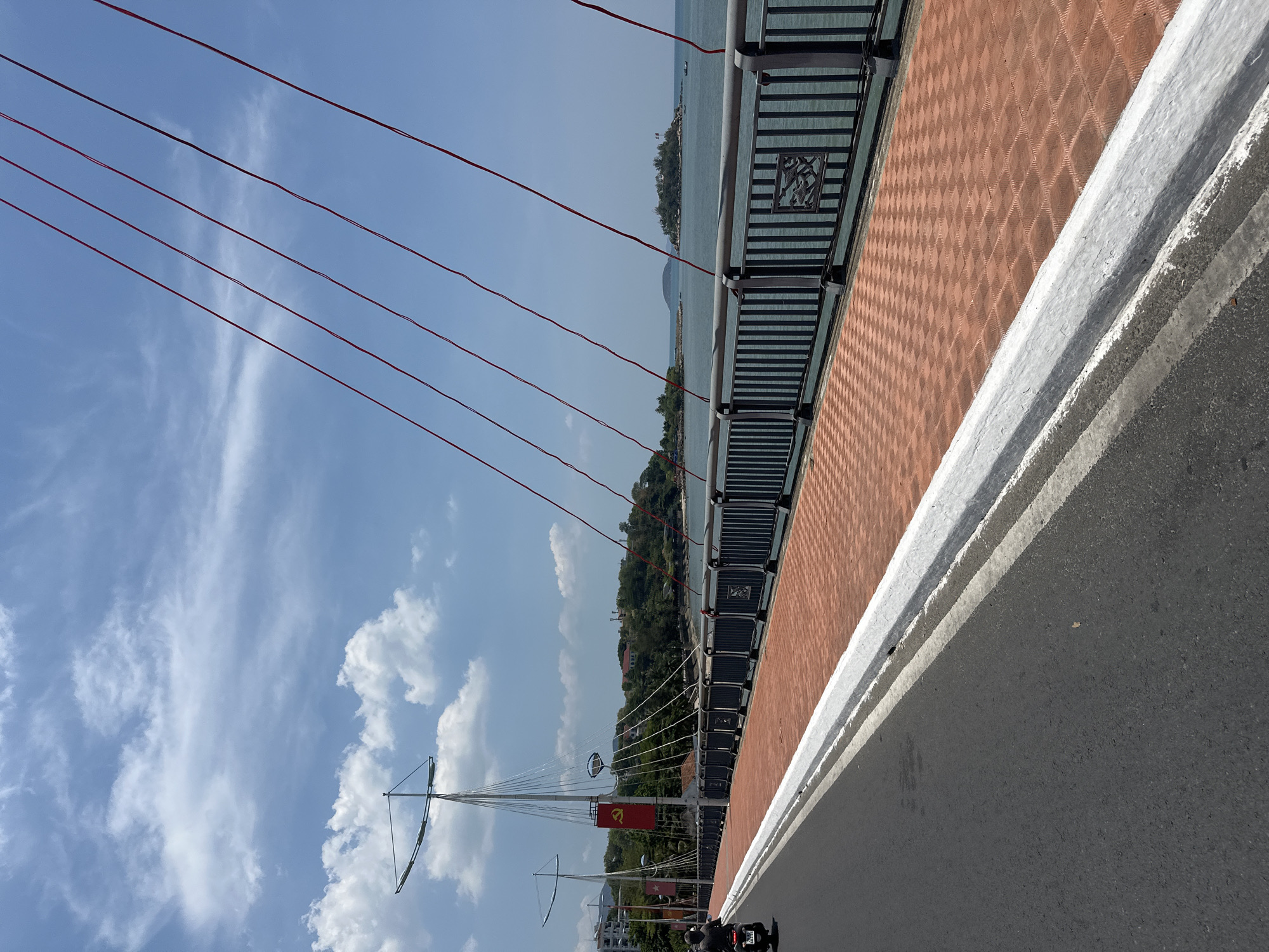 The image shows a view from a bridge in the coastal city of Bac Nha Trang Ward, Vietnam. The bridge has a brown and black patterned barrier. Red cables span across the sky, which is partially cloudy. A red flag with a yellow star and hammer/sickle emblem is visible on a light pole extending from the bridge. A stretch of water and a distant island are visible behind a tree line. The bridge has a gray road surface with a white line marking the edge. A motorcycle is parked at the end of the bridge.