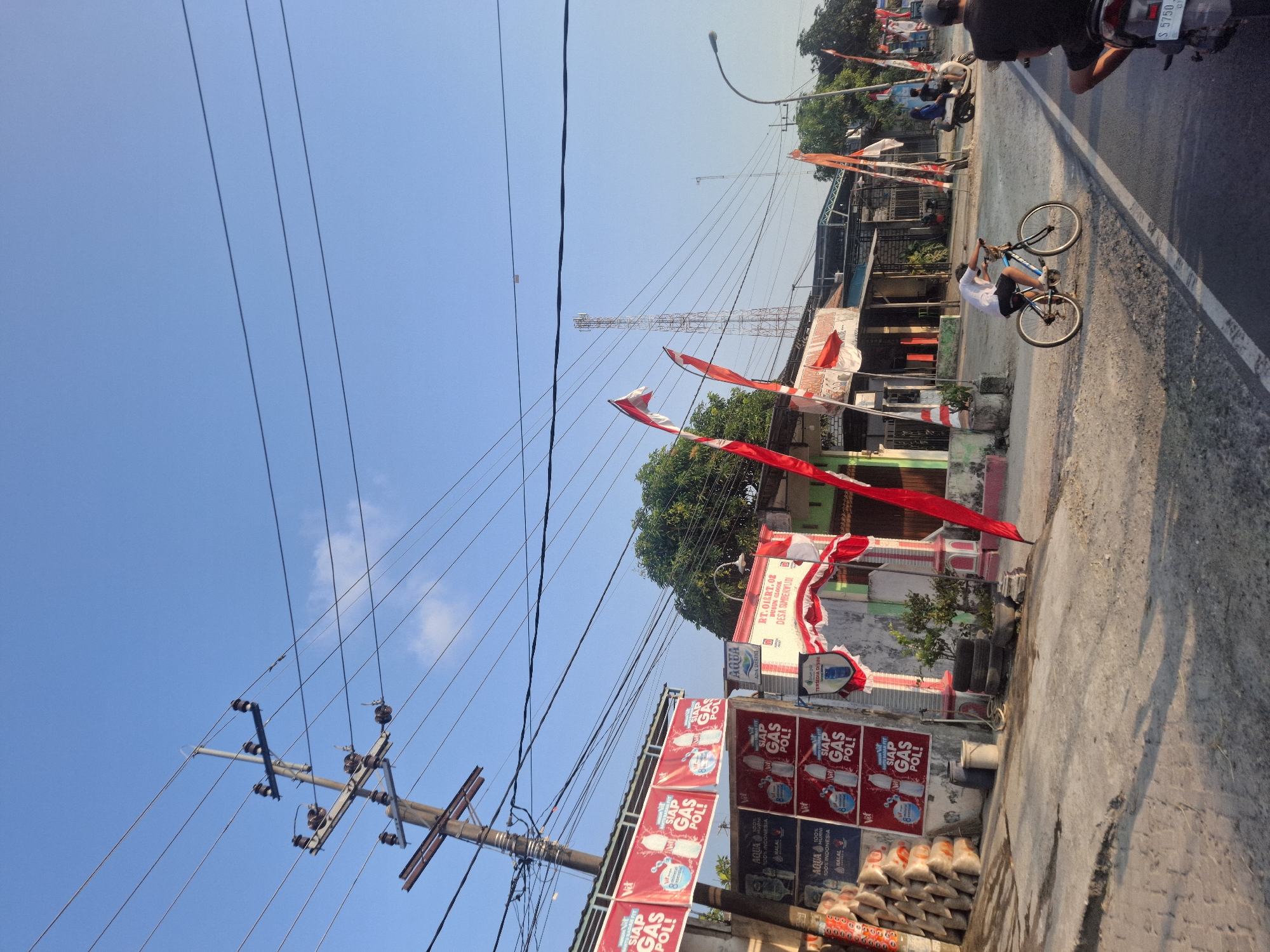 A street scene in Glogok, Indonesia, under a clear blue sky features a utility pole with multiple wires on the left, and several Indonesian flags (red and white) are displayed along the road. Several shops or stalls are visible on the right side of the street, adorned with red and white banners. A young person is riding a bicycle on the dirt shoulder of the road, while a motorcycle with two occupants is further down the street. The ground is paved in some areas and dirt in others, with shadows cast by the overhead wires and structures. Several stacks of bags and tires are also present near the buildings.