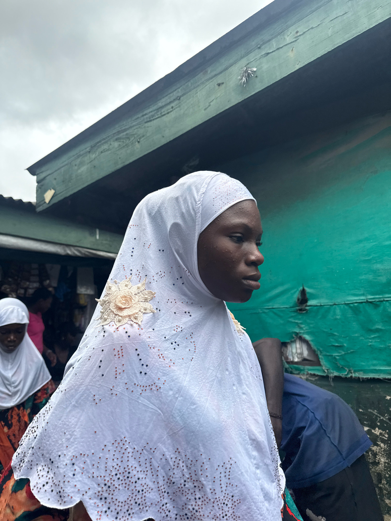 An outdoor, medium shot captures a young woman in a white hijab with intricate beadwork and a floral embellishment, standing in profile and looking to her right. She is positioned in the foreground, obscuring a portion of her body and face. Behind her, slightly out of focus, another young woman in a similar white hijab is visible, wearing a colorful garment. In the background, a man in a navy blue shirt and dark pants sits, his arm resting on something unseen. The environment appears to be an urban setting in Lagos, Nigeria, with a weathered, green, corrugated metal structure overhead, suggesting a market or street stall. The sky is overcast and grey, and the overall lighting is dim. The scene implies a candid moment in a bustling, possibly underprivileged, part of the city.
