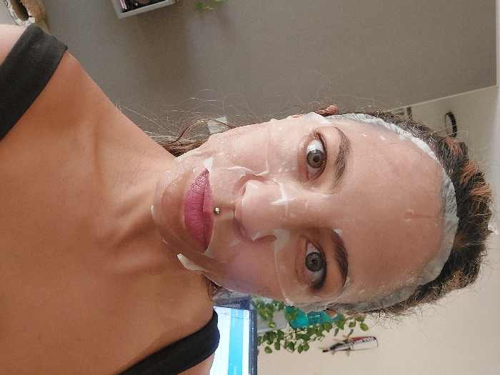 The image displays an adult female indoors, captured in a mid-shot with her face directed towards the viewer. A translucent sheet mask covers her entire face, featuring cut-outs for her eyes, nostrils, and mouth, with visible liquid residue. Her eyes, appearing green or hazel, are open and focused on the camera. A silver labret piercing is positioned centrally above her upper lip, which exhibits a pinkish tint. Her hair is pulled back from her face. She wears a black garment with a thin strap visible over her right shoulder and upper chest. The background on the left is a plain light grey wall, with a partial dark circular object (possibly a light fixture) visible near the top left. On the right, a lighter-toned wall features a hanging green plant. Below the plant, a portion of a computer monitor screen displaying a light-colored interface is visible, alongside another indistinct object. The specific location within Buenos Aires, Argentina, cannot be visually identified from the scene.