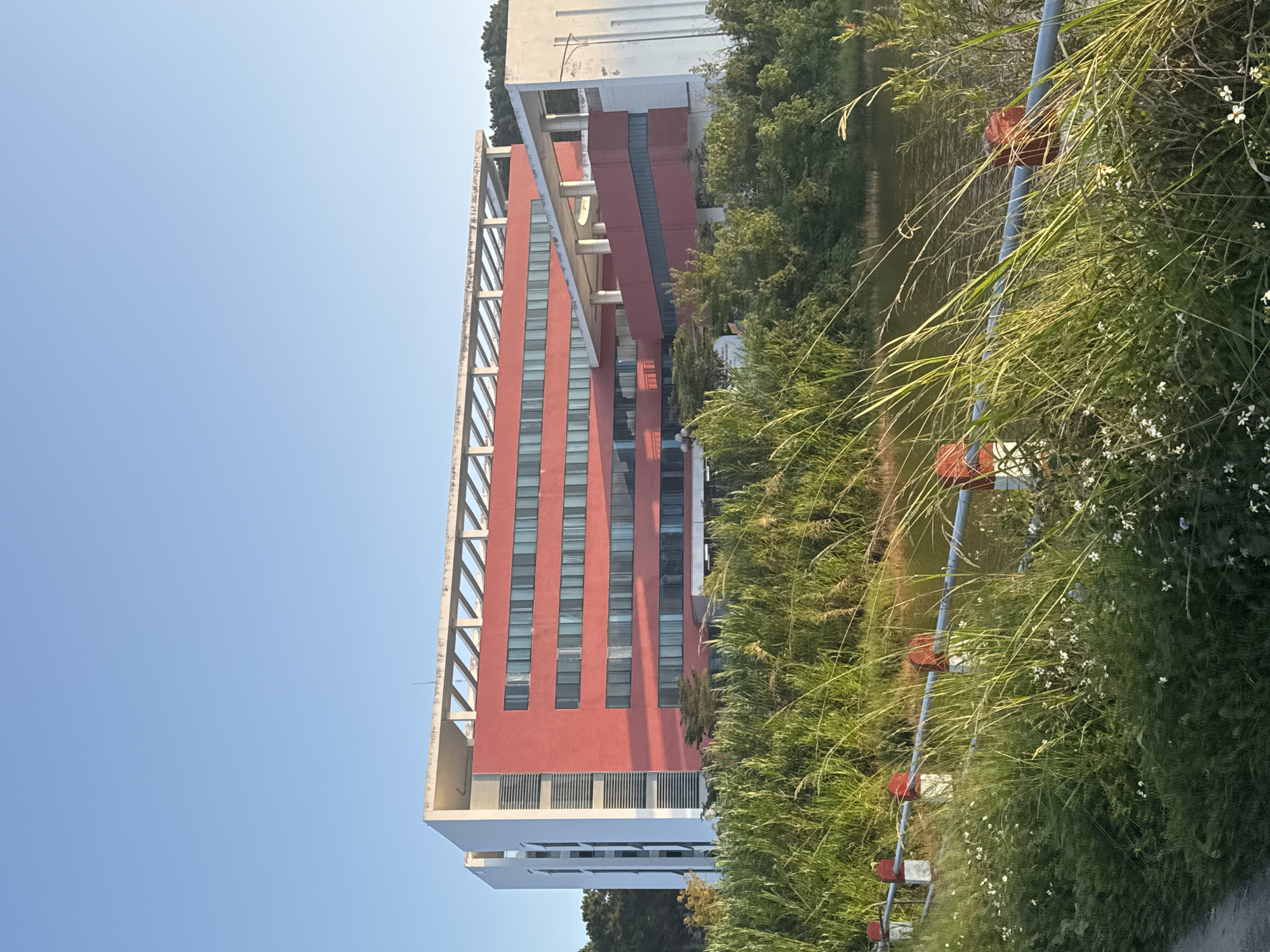 This image shows a multi-story building with a red facade and white accents, located in Lâm Viên - Đà Lạt, Vietnam. The building is situated behind a body of water and a grassy embankment with a metal railing. Tall green grass and some small white flowers are in the foreground, partially obscuring the view of the railing and the water. The sky is clear and blue, indicating it is daytime. The building's architecture features numerous windows and a distinctive latticed overhang at the top. The overall scene suggests a tranquil setting, likely on the outskirts of a city or within a campus environment.