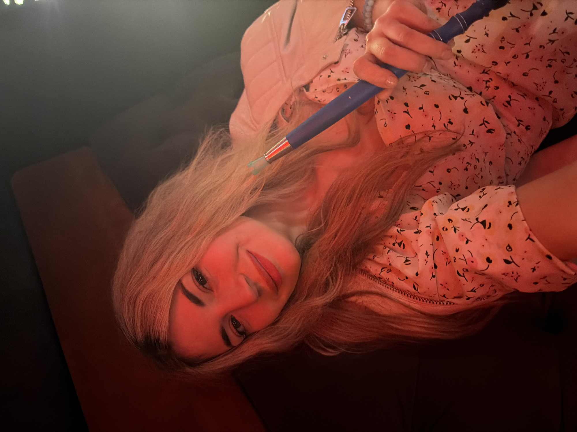 A woman with long blonde hair is holding a blue hookah mouthpiece to her lips. She is wearing a white floral top and a light-colored jacket. The lighting is dim and reddish. The background appears to be a dark-colored couch or seating area. The image does not contain information to specify the city of Budva, Montenegro.