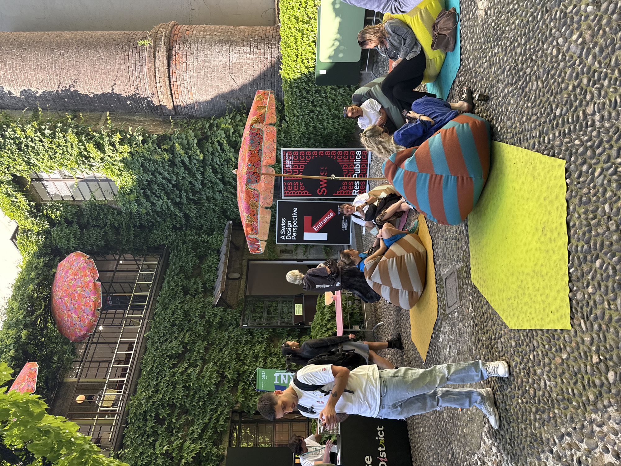 The image depicts an outdoor gathering in Milan, Italy. People are seated on colorful mats and beanbags on a cobblestone surface. Promotional materials, including a sign reading "A Swiss Design Perspective" and "Res Publica," are visible. The event is sponsored by Audi, indicated by a banner. A man in a white shirt and light jeans stands in the foreground, looking at his phone. The buildings are covered in ivy, with patterned umbrellas providing shade. A large brick structure is seen in the background.