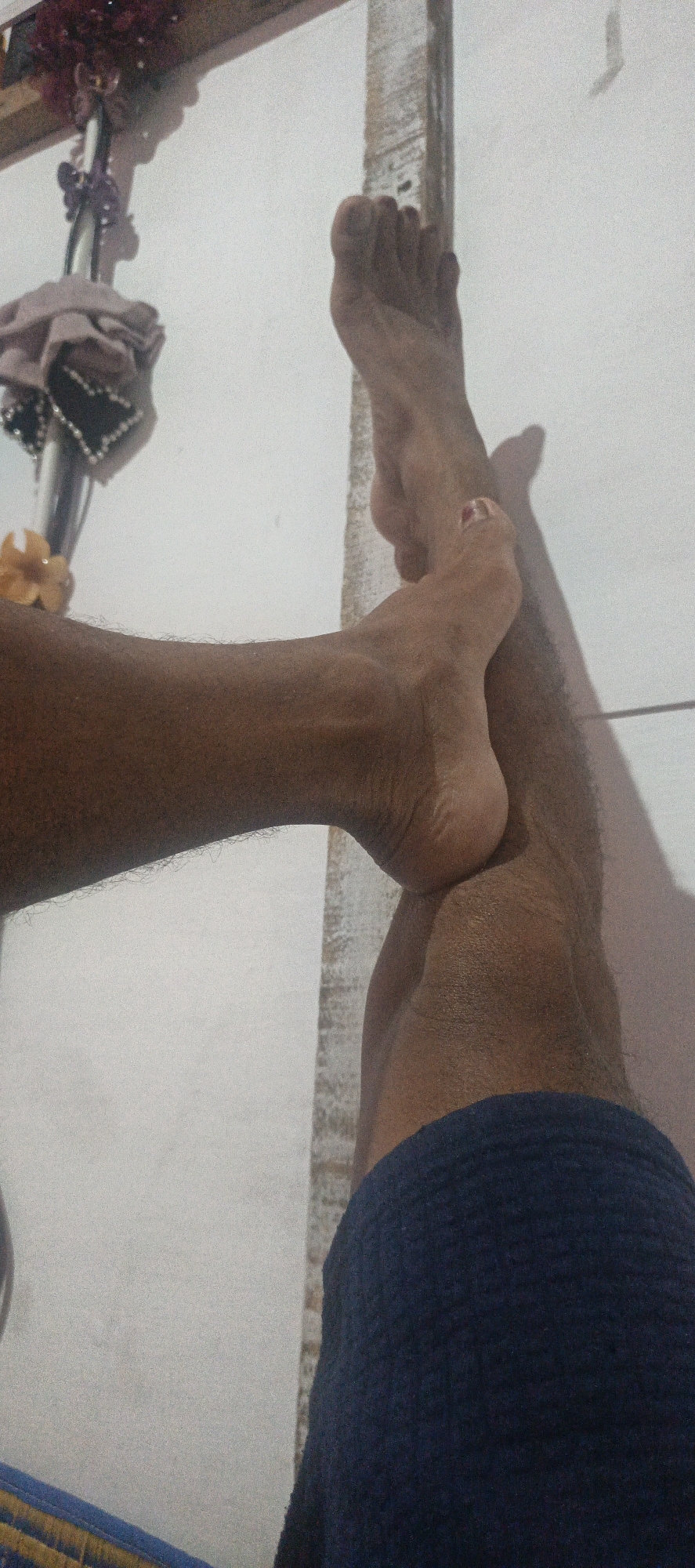 The image displays a close-up view of two bare feet and lower legs against a textured white wall. The feet and legs are tanned, with visible hair on the legs. One foot is positioned on top of the other, with the toes of the upper foot pointing upwards. The toes of the lower foot are visible and appear to have a small amount of nail polish on one toe.
In the background, to the left, there is a decorative object with red flowers and some fabric draped over it, attached to a vertical pole. The wall appears to be a simple indoor surface. The lighting creates a shadow of the legs and feet on the wall. The bottom of the image shows a glimpse of a dark blue garment, possibly shorts or a skirt. The scene is devoid of any context that would suggest a specific location within Cinta Dame, Indonesia, or any particular activity or interaction beyond the arrangement of the feet and legs.