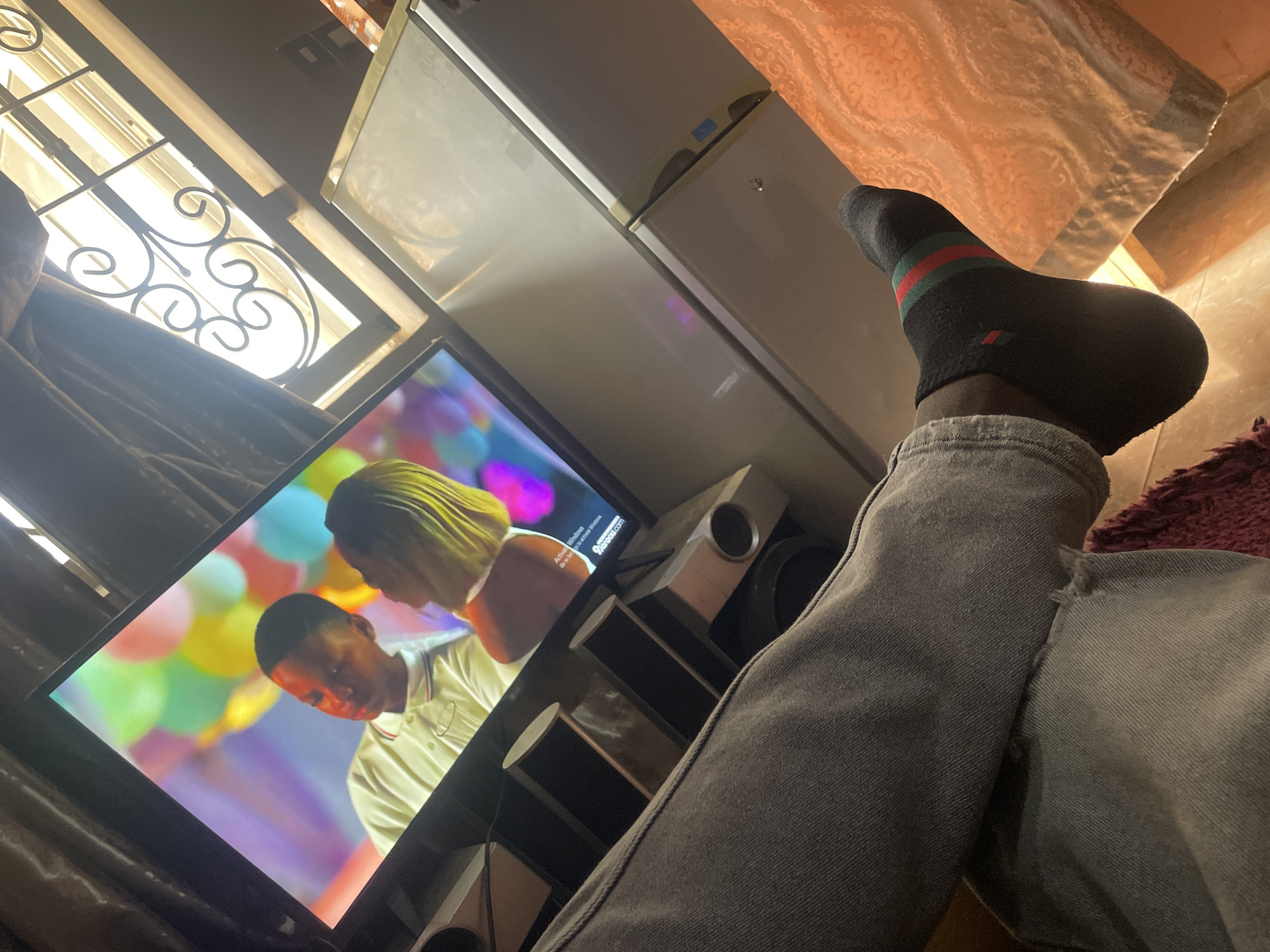 A person wearing grey jeans and a black sock with red and green stripes is watching a television. The television is showing a scene with a man and a woman, surrounded by colorful balloons. Adjacent to the television are several speaker towers. To the side of the television and speakers is a refrigerator. A window with decorative wrought iron is visible in the upper left corner of the image. Based on the prompt, the location is assumed to be Jos, Nigeria, but there is no direct visual evidence to confirm this location.