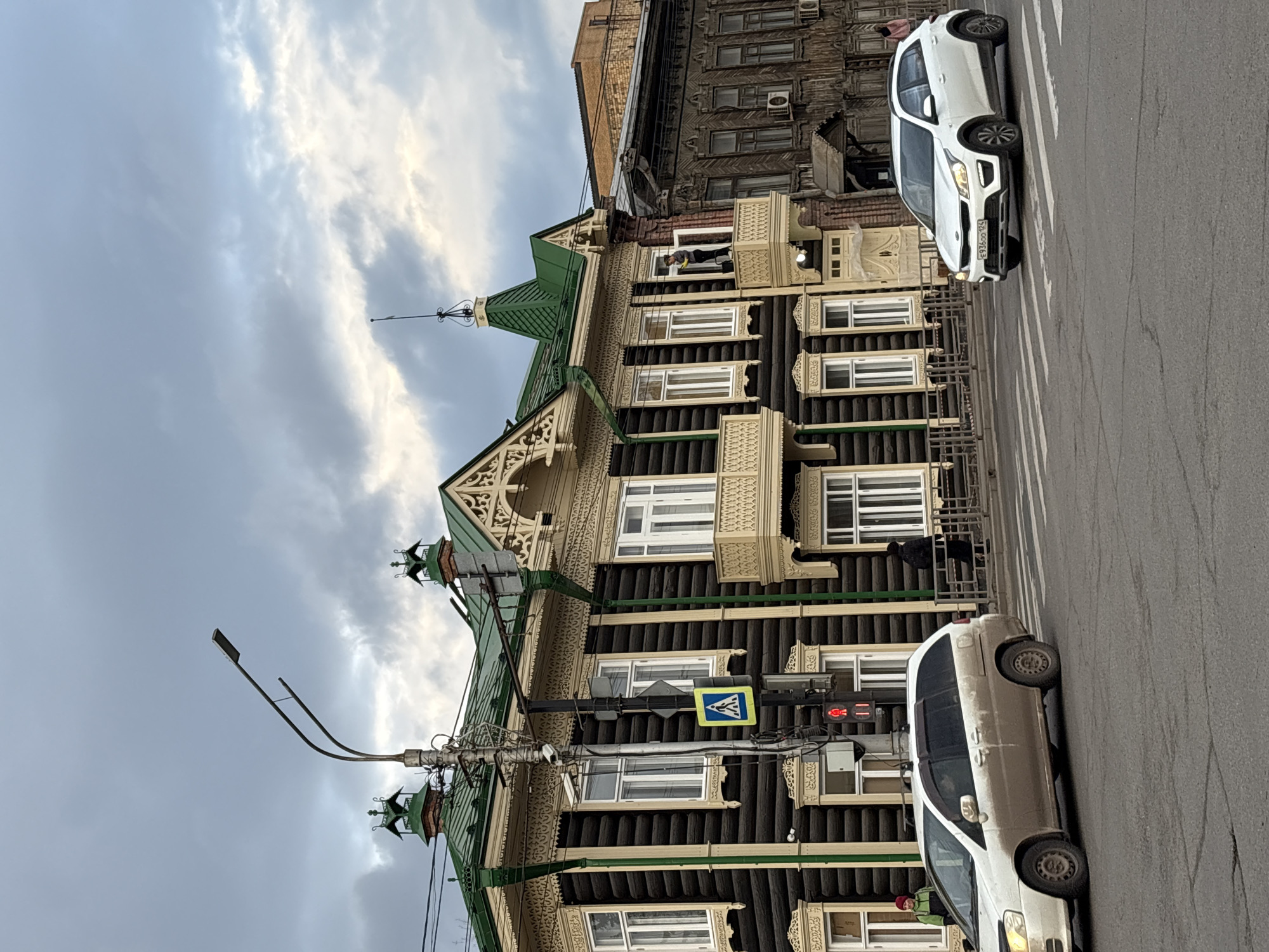 A street scene in Krasnoyarsk, Russia. A two-story wooden building with intricate carved details and a green roof is positioned beside the street. The building has a pedestrian crossing sign and a traffic light in front of it that is red. Two white cars are on the road, one in the foreground and the other in the background. The sky is overcast. Another building is visible to the right.