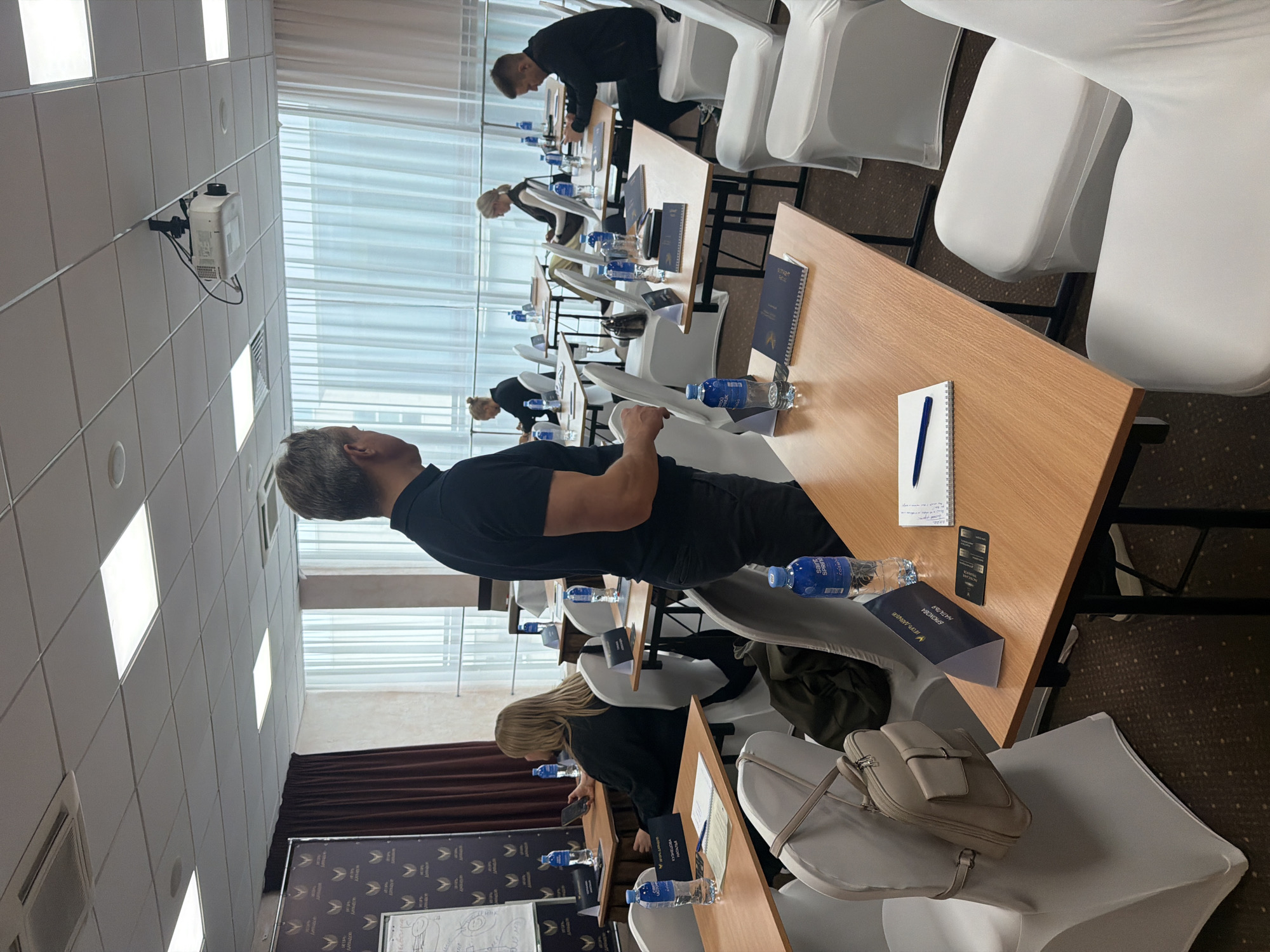 The image shows a classroom or conference room in Pervomaysky, Russia, with several rows of wooden tables covered in study materials. People, mostly men, are seated in white chairs around the tables, seemingly engaged in a lecture or presentation. Notebooks, pens, water bottles, and branded materials are on the tables. A projection screen is set up at the front of the room, and the windows are covered with semi-transparent blinds. A projector is affixed to the ceiling. The overall environment suggests a formal educational or training setting.
