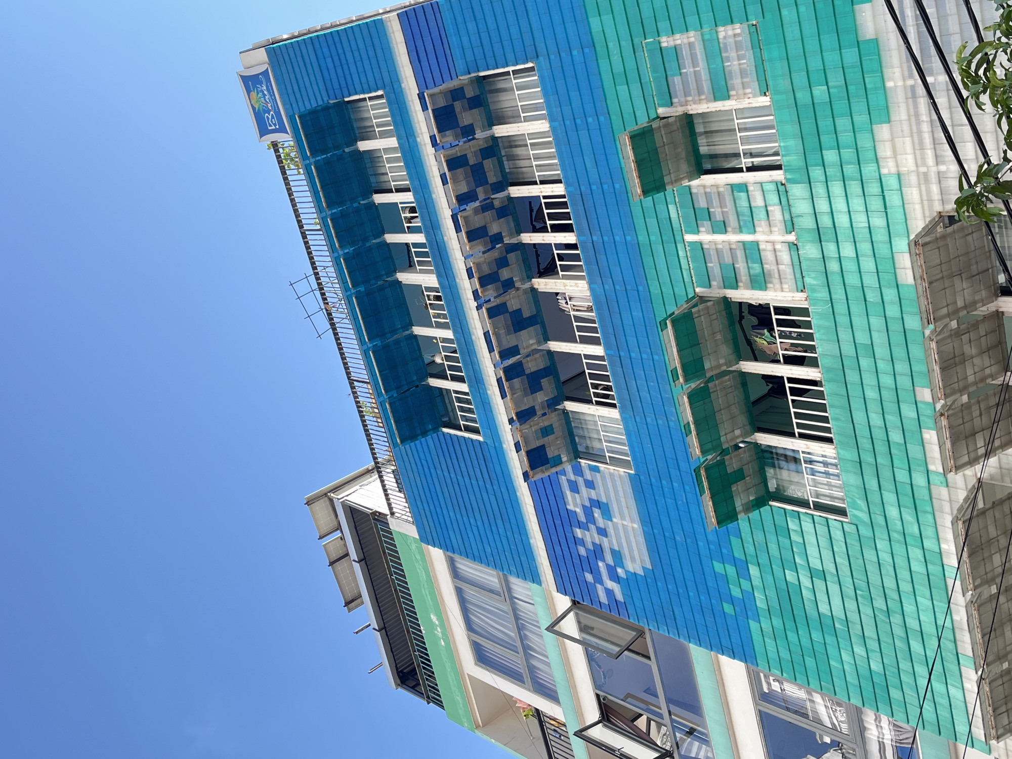 A low-angle shot captures a building with a colorful facade under a clear blue sky. The building features alternating blue and green vertical panels, with rows of windows visible on each floor. Some windows are covered with netting or have awnings, while others are open. A balcony with a railing is seen on the upper left side of the building. A sign is visible near the top left corner. Power lines are visible against the building.
The building is located in Đà Nẵng, Vietnam.
