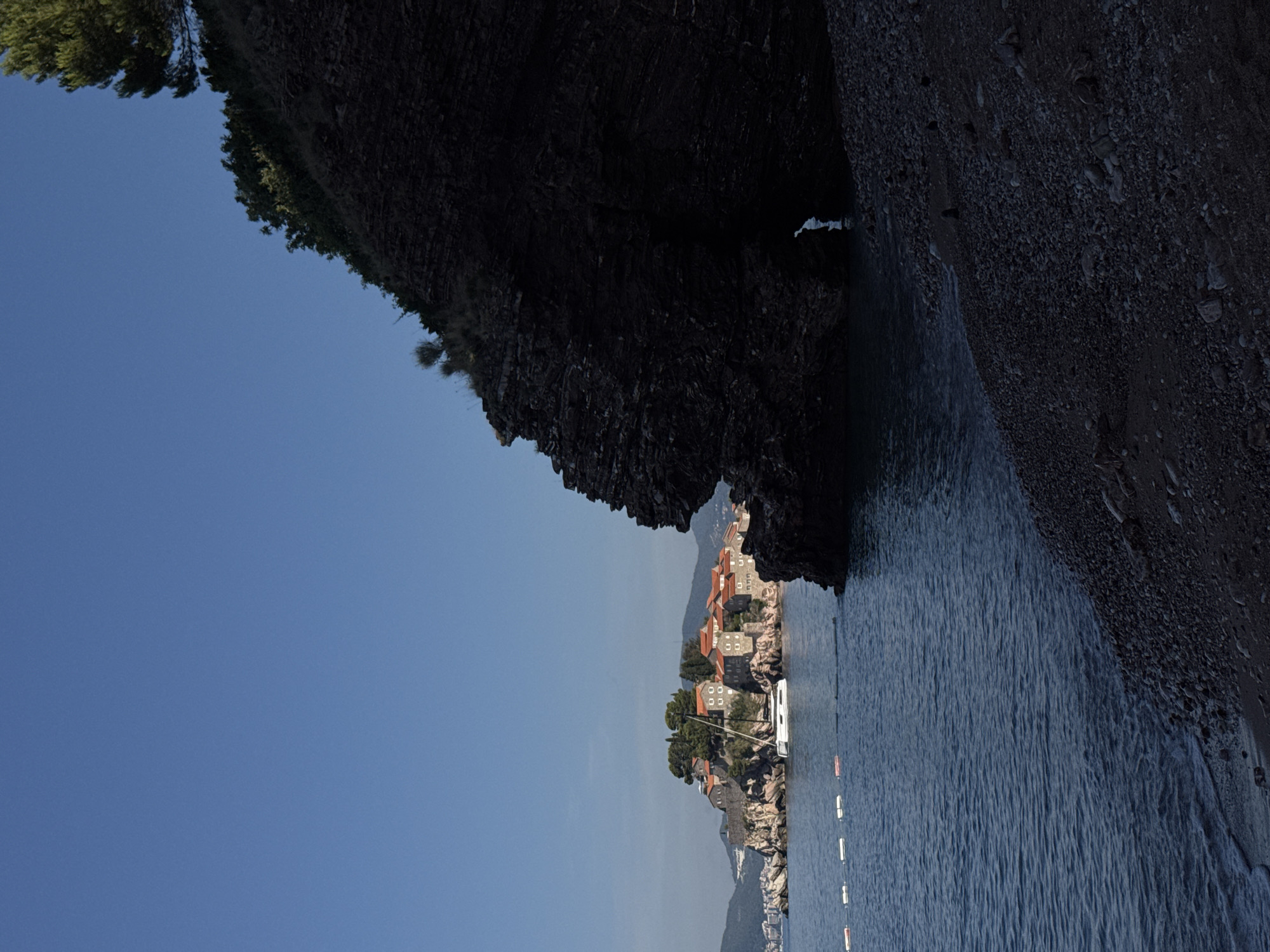 The image depicts a scenic view of the island of Sveti Stefan, Montenegro, from a rocky shoreline. In the distance, the historic island is densely populated with red-roofed buildings and lush greenery, connected to the mainland by a narrow causeway which is not visible in this frame. A white sailboat is anchored in the calm, blue waters between the island and the foreground. The foreground is dominated by a large, dark, rocky cliff formation with a visible opening or cave leading to the water. The beach in the foreground is a mix of sand and dark pebbles, with small waves lapping at the shore. A line of white buoys with orange accents floats on the water, demarcating a boundary. The sky is clear and blue, indicating a sunny day. There are no people visible in the frame.