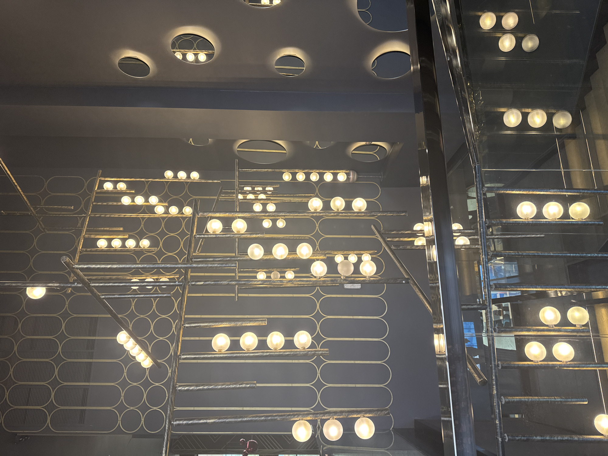 The image depicts the interior of a modern building, possibly a hotel or a restaurant, with a focus on a striking chandelier and a staircase. The chandelier features numerous clear glass globes emitting a warm light, suspended at varying heights by metallic rods and cables. The rods are part of a larger, intricate metal structure that also serves as a railing for the staircase. The walls are painted a dark gray, and decorative circular mirrors with a soft glow are embedded in the wall on the right side of the image, complementing the lighting. The staircase itself is also metallic, with steps visible in the lower portion of the frame. The overall aesthetic is contemporary and sophisticated, with a strong emphasis on lighting design. The scene is devoid of people. There is no visible indication of the city being Moscow, Russia.