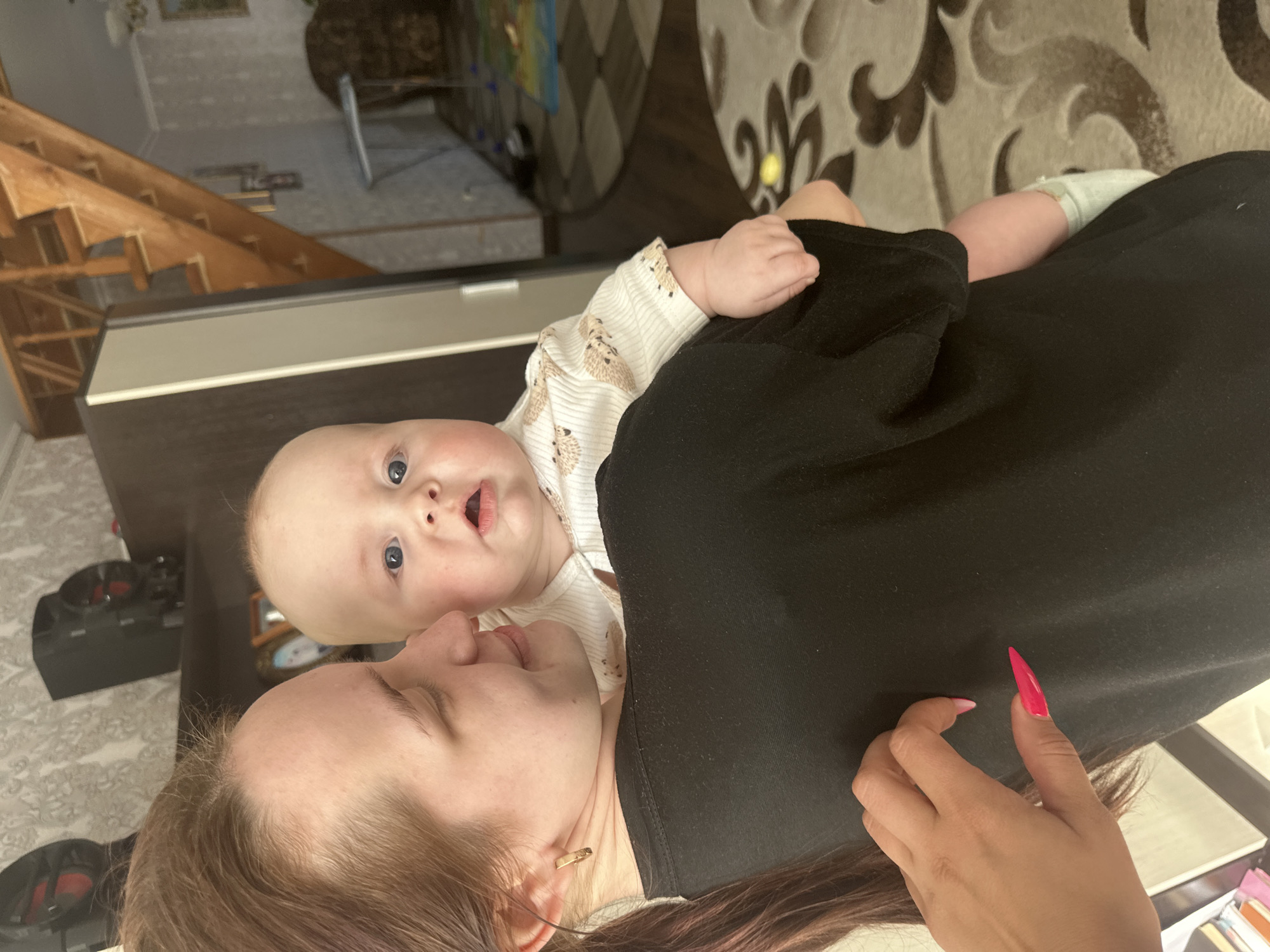 The image, rotated 90 degrees clockwise, captures a woman holding a baby indoors. The woman, with light brown hair pulled back, is wearing a plain black top and a small gold stud earring in her left ear. Her left hand is visible, featuring long, sharply pointed bright pink stiletto nails. She is looking downwards and to the left, towards the baby, with a soft expression.
The baby, with fair skin and striking light blue eyes, is looking directly at the camera with a wide-eyed, curious or surprised expression, mouth slightly agape. The baby is dressed in a white long-sleeved onesie patterned with small brown hedgehogs. One of the baby's small hands is resting on the woman's black top, while a foot, partially clad in a light blue sock, peeks out near the woman's side.
The background suggests a home environment. A dark, flat surface, possibly a counter or piece of furniture, is directly behind the woman and baby. On the far left, a wooden staircase or complex shelving unit is visible. The floor is covered by a patterned rug featuring large, swirling designs in shades of brown and beige. Another dark piece of furniture can be seen further in the background. The lighting is diffused and even, indicating an indoor setting. No text is visible in the image. The scene portrays a tender interaction between the two individuals.