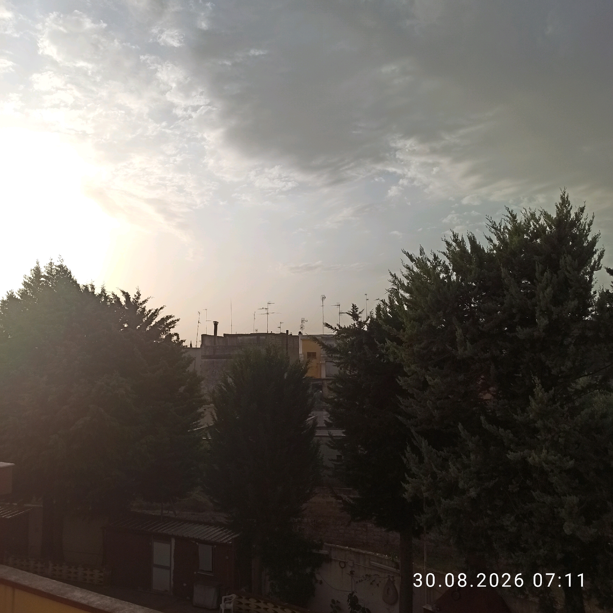 The image captures a view of the sky over the city of Bitonto, Italy. The sky is filled with clouds, with a bright sun visible on the left side, partially obscured by clouds. In the foreground, there are several evergreen trees, some of which are silhouetted against the bright sky. Behind the trees, a cityscape is visible, with rooftops, buildings, and numerous television antennas. The scene is set during the day, as indicated by the lighting and the time stamp "30.08.2026 07:11" in the lower right corner. The overall impression is that of an urban landscape viewed from an elevated position, possibly a balcony or rooftop, looking towards the sky and the surrounding buildings. There are no people visible in the image.