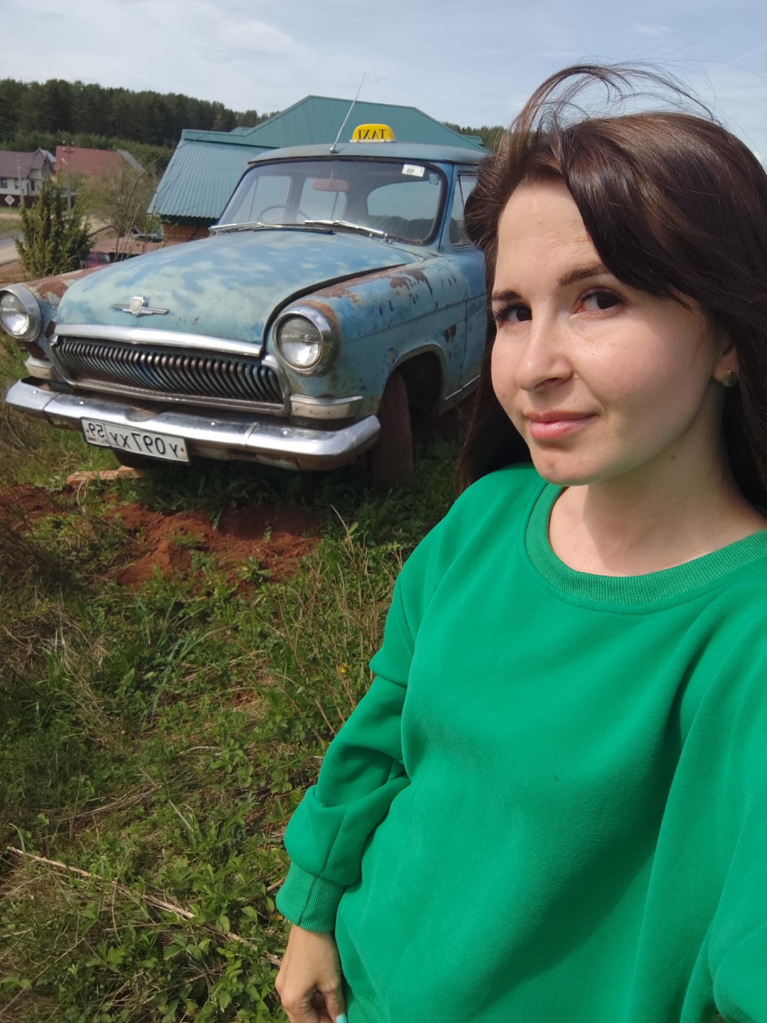 A young woman wearing a green sweatshirt stands outside in a grassy area in Фоки, Russia. Behind her is a weathered, light-blue vintage car with a "TAXI" sign on its roof, indicating it was formerly used as a taxi. The car is positioned on a mound of dirt in the grass. The environment includes a building with a green roof and other structures visible in the background.